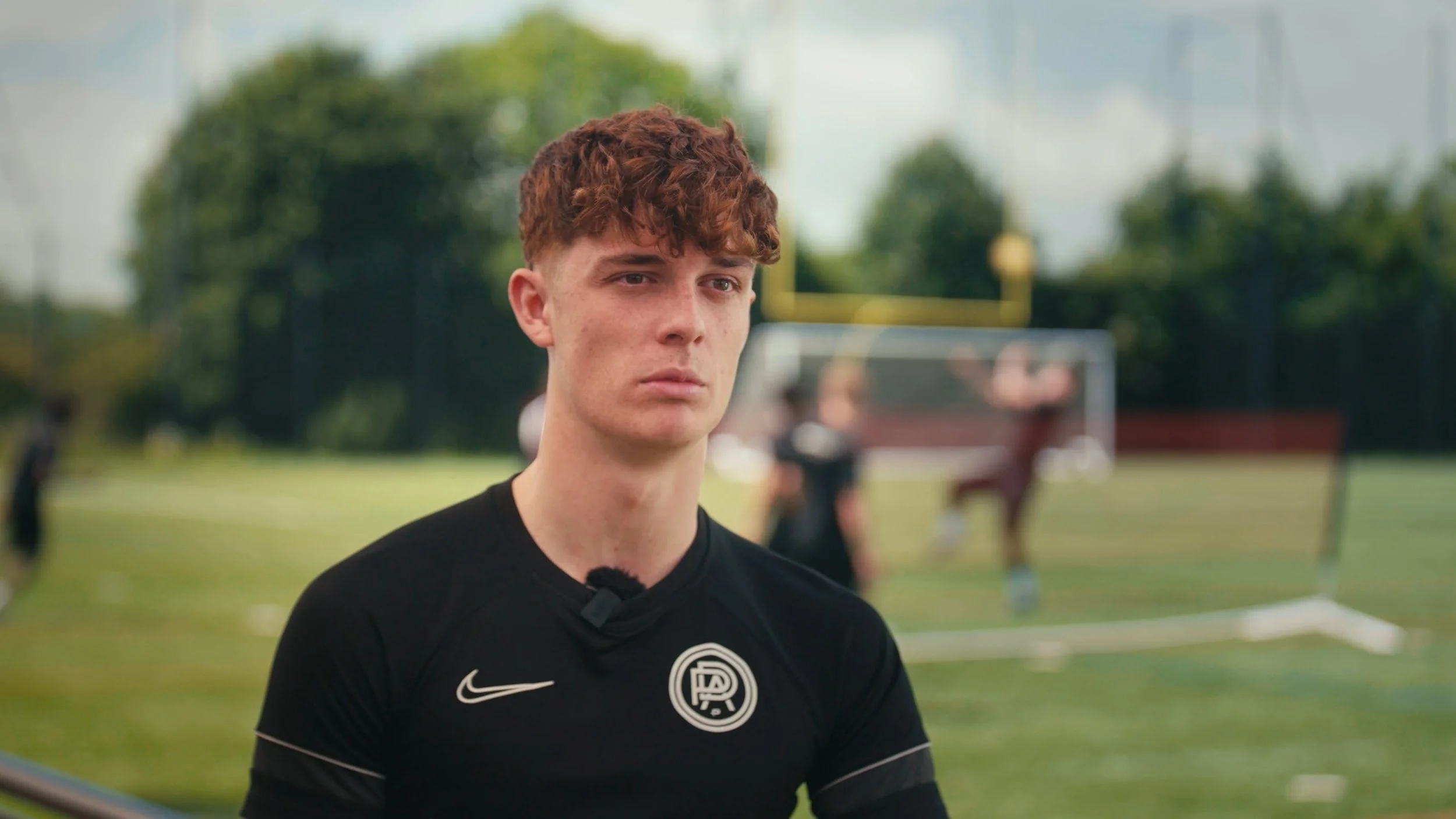 A young man with curly red hair wearing a black sports jersey sits on a football field during an interview, with a goal net and other players practicing in the background.