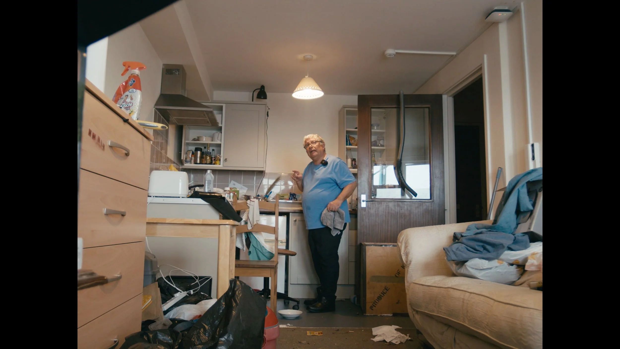 A cluttered kitchen with a woman in a blue shirt standing near the counter, surrounded by various items and clutter, including a sofa with laundry on it, a table with items, and trash on the floor.