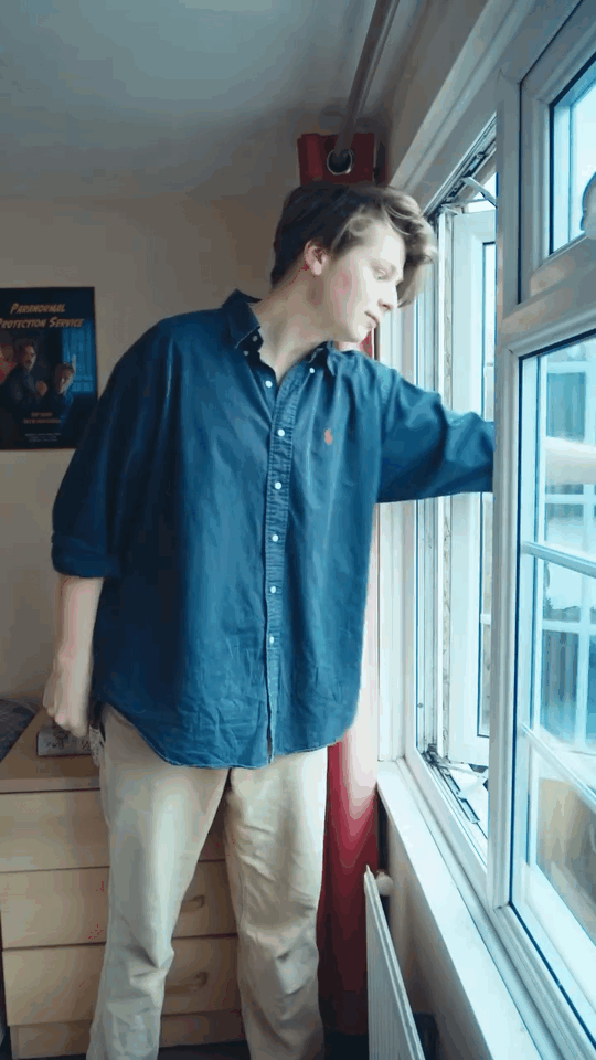 A young man with brown hair wearing a dark blue button-up shirt and beige pants, standing by a window looking outside.
