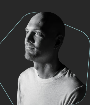 A man in a red sports jacket sitting in a room with a display case of trophies and a large screen showing logos.