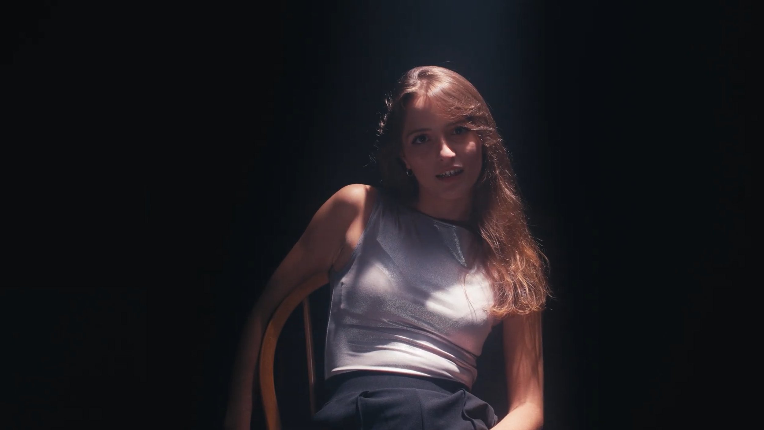 A woman with long, wavy brown hair sits in a dark room, illuminated by a single overhead light. She is wearing a sleeveless satin top and dark pants, looking at the camera with a neutral expression.