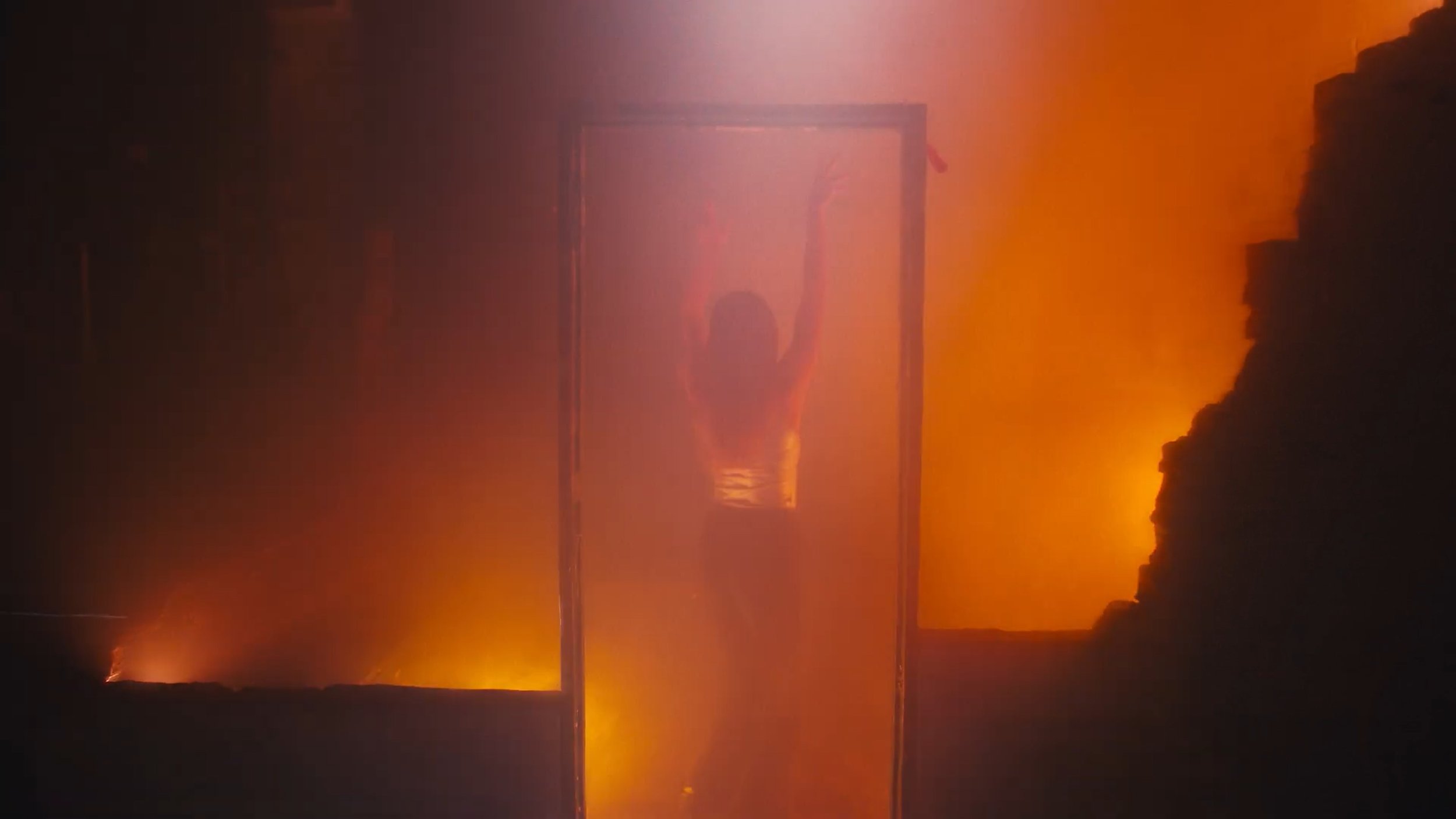 Person standing in a doorway with fire and smoke behind them, casting an orange glow in the scene.