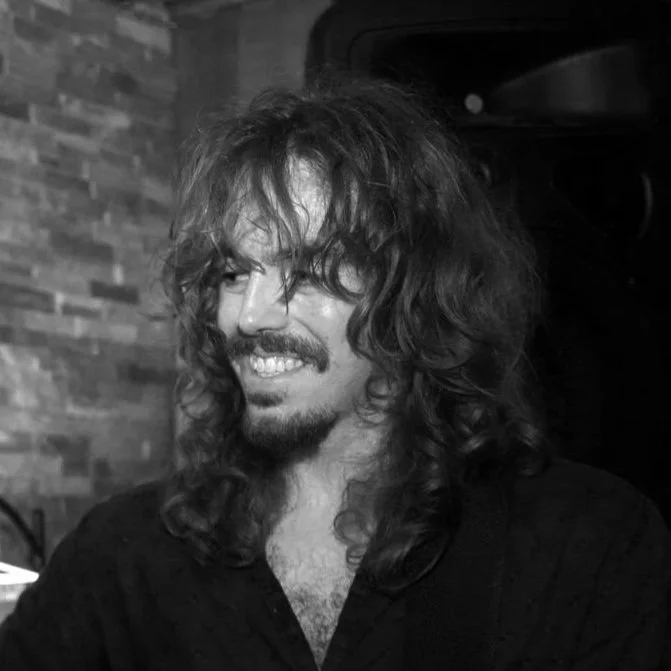 A man with long, curly hair and a beard smiling in a black and white photo.