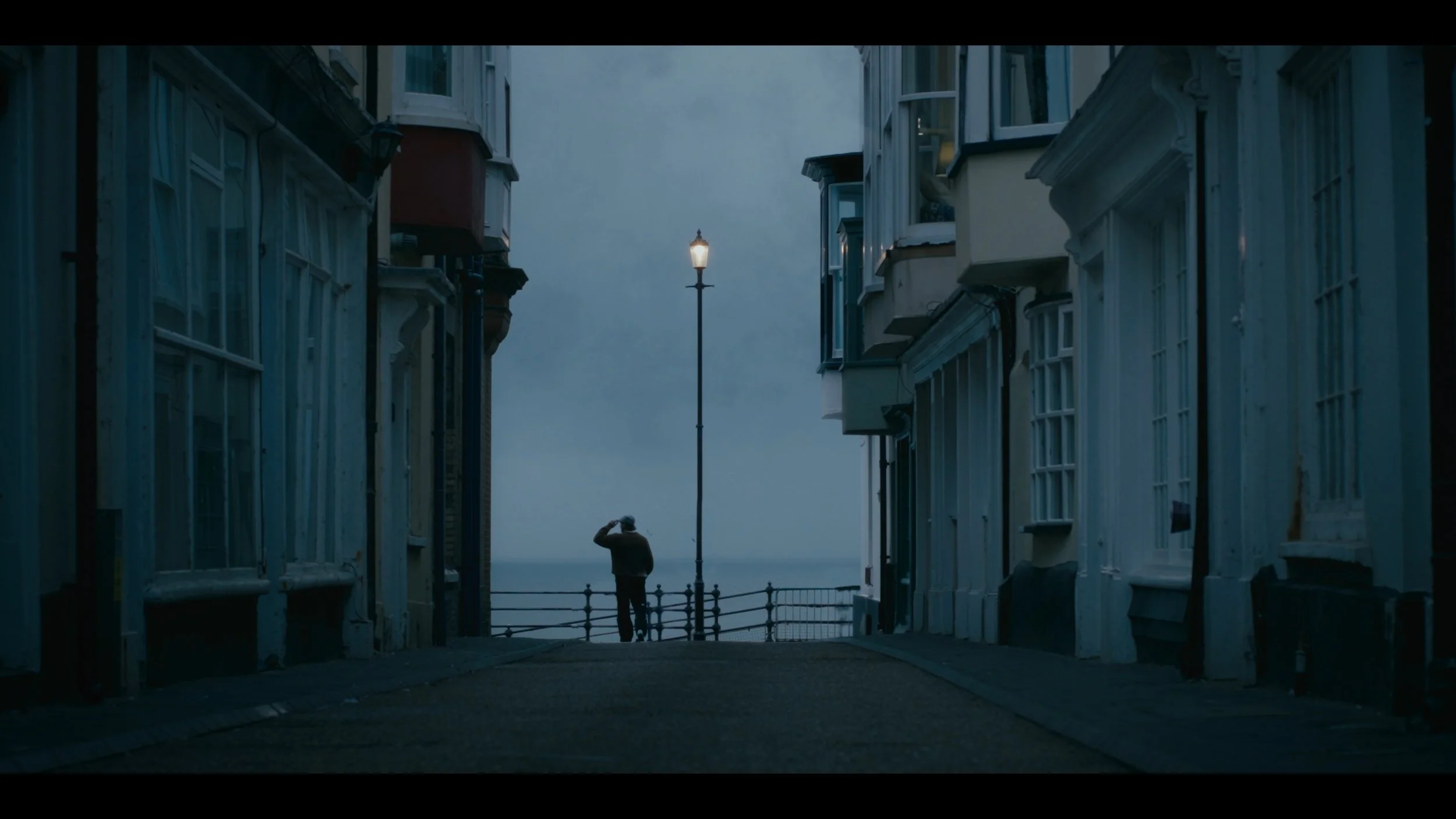 A person standing on a street near the coast, looking out over the water, with a lamppost in the foreground amid close-up buildings on either side.