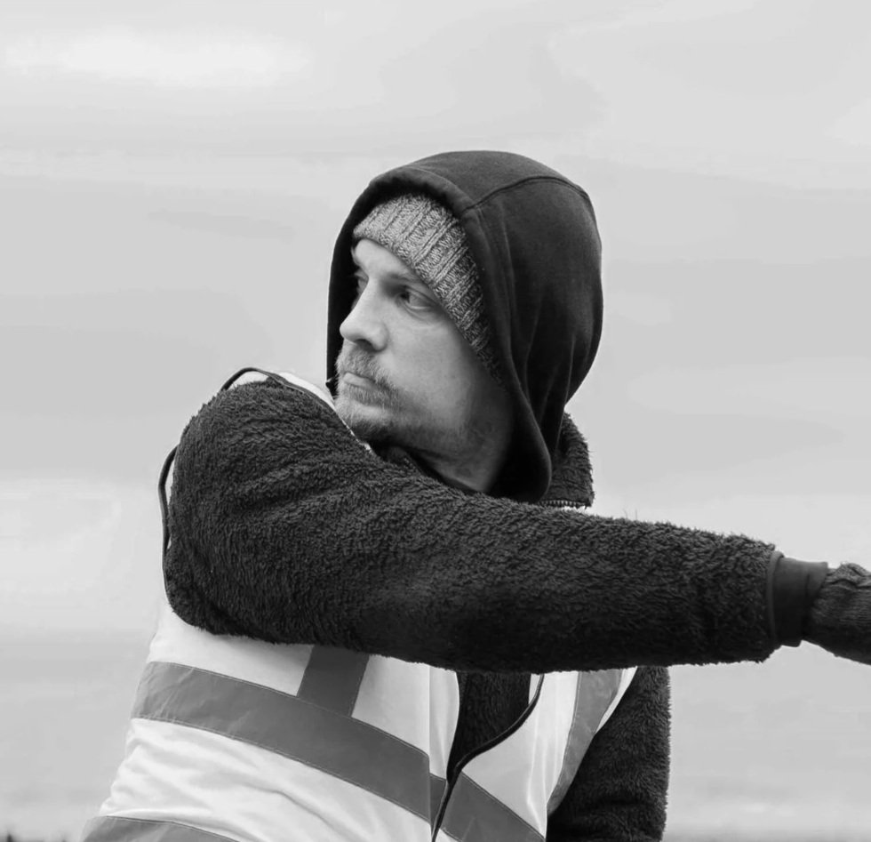 A man wearing a hoodie, beanie, and jacket, looking to the side with an arm extended forward.