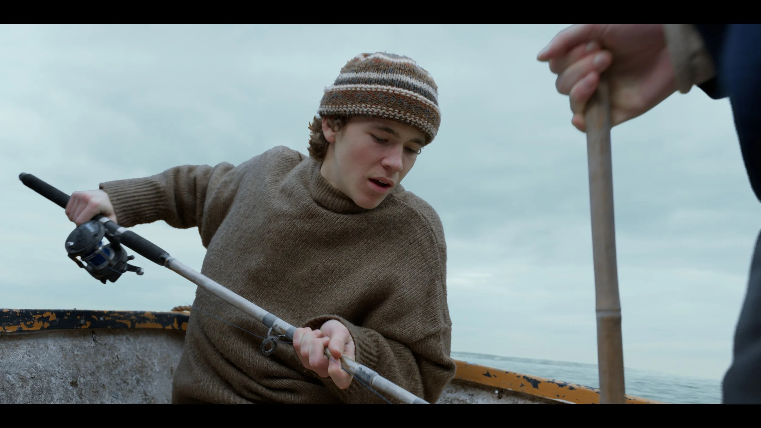 Young man in a brown sweater wearing a knit cap holding a fishing rod on a boat, with another person holding a bamboo pole nearby, over a body of water.