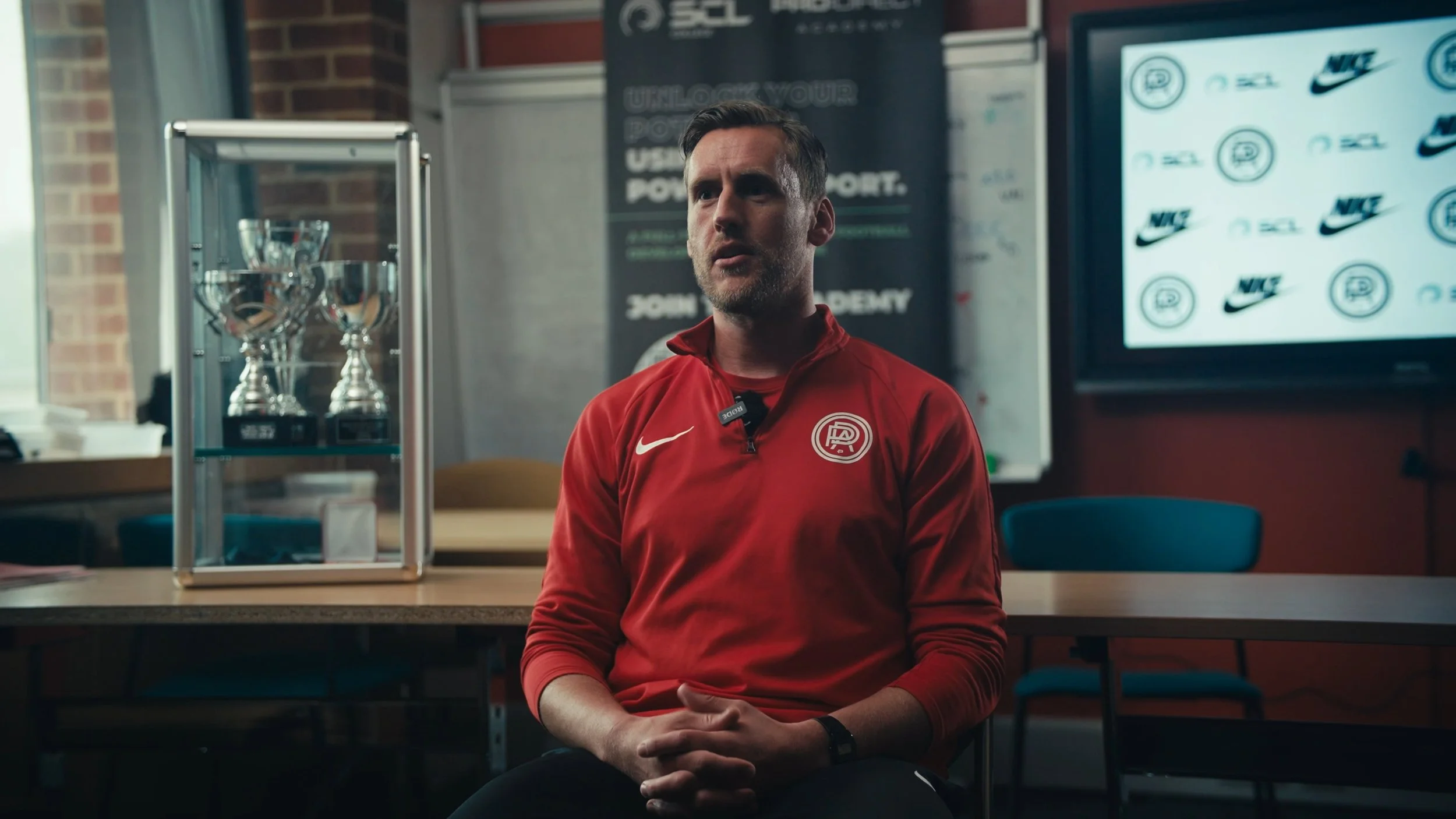 A man wearing a red sports jacket with a logo on the chest sits in a room with a black background, a monitor with sports logos, glass trophies on a display case, and a poster in the background.