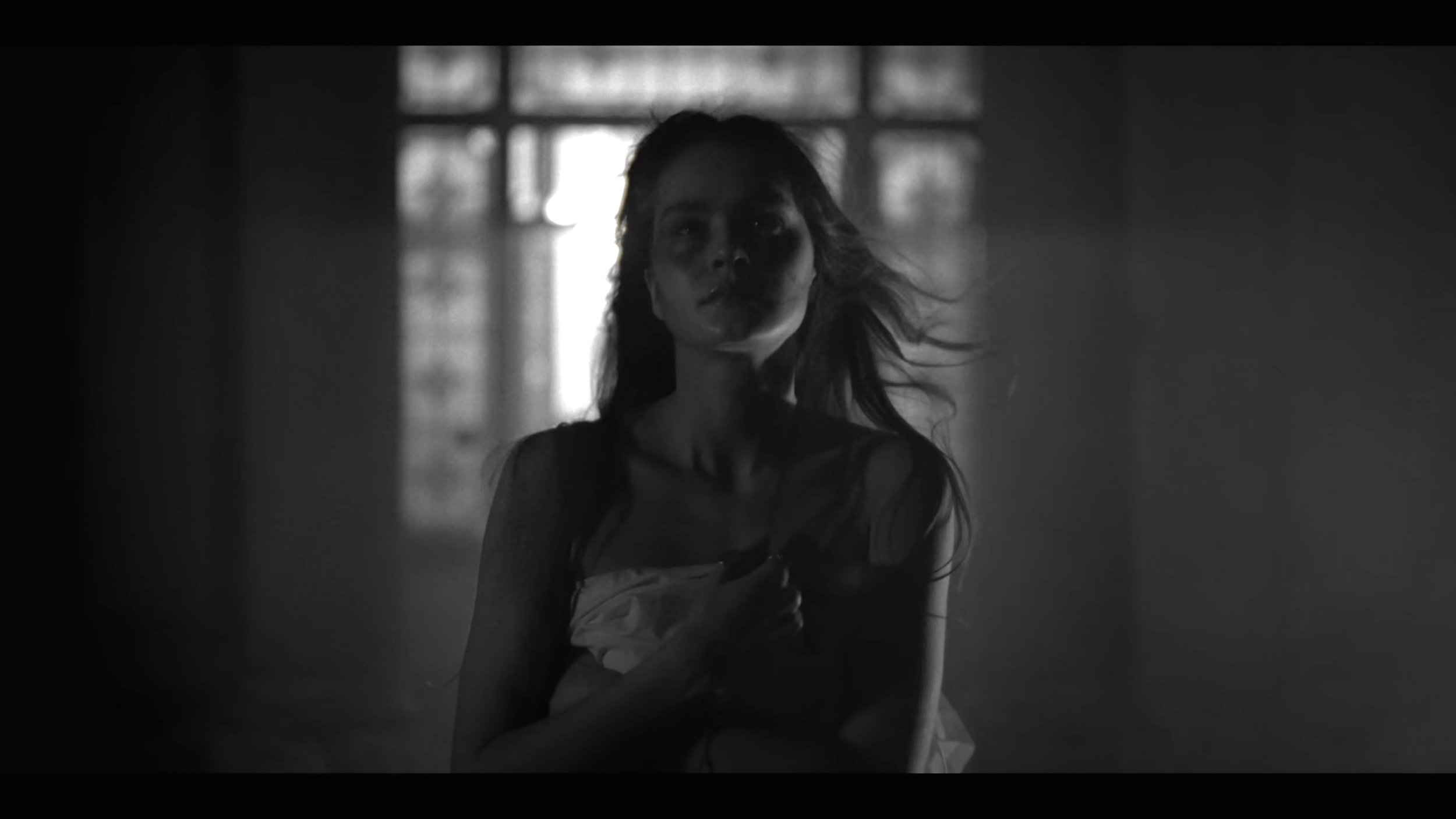 Black and white image of a woman with disheveled hair standing in front of a window, holding her shirt to her chest, with a somber expression.