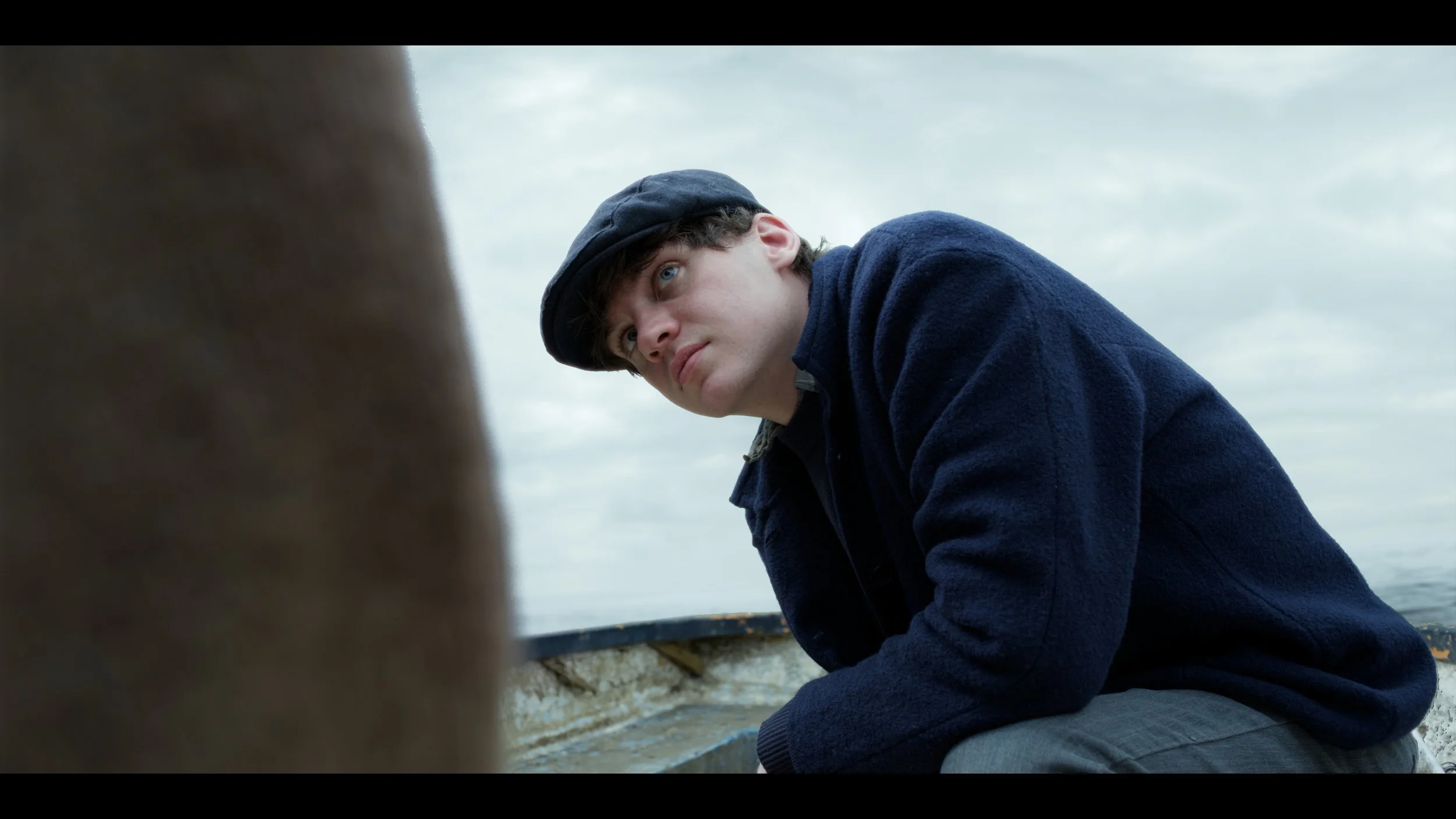 A young man wearing a navy blue sweater and navy cap, sitting in a boat on the water, looking serious or contemplative.