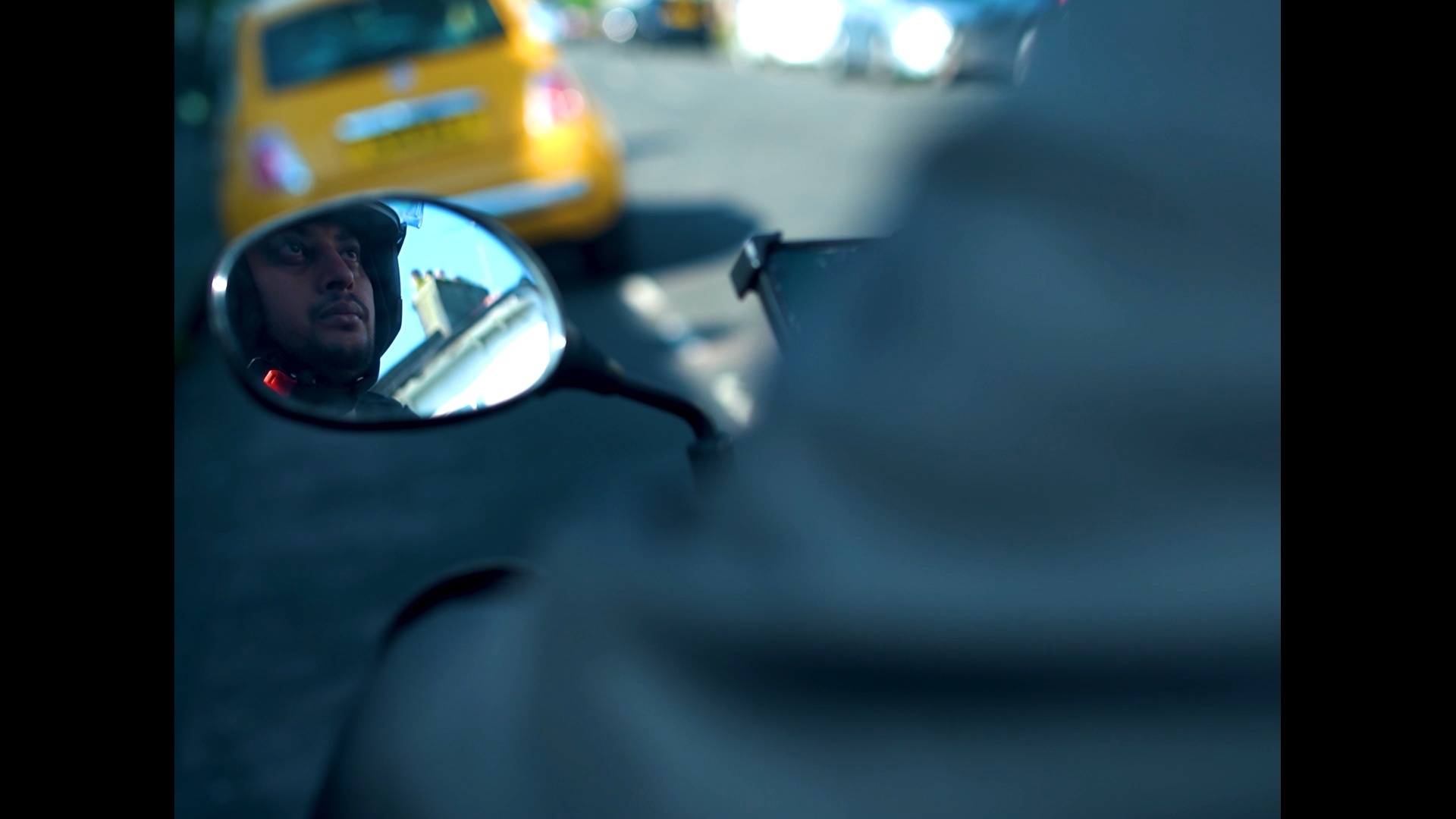 Close-up view from inside a vehicle showing a side mirror reflection of a person wearing a helmet, with a yellow car and cityscape in the background.