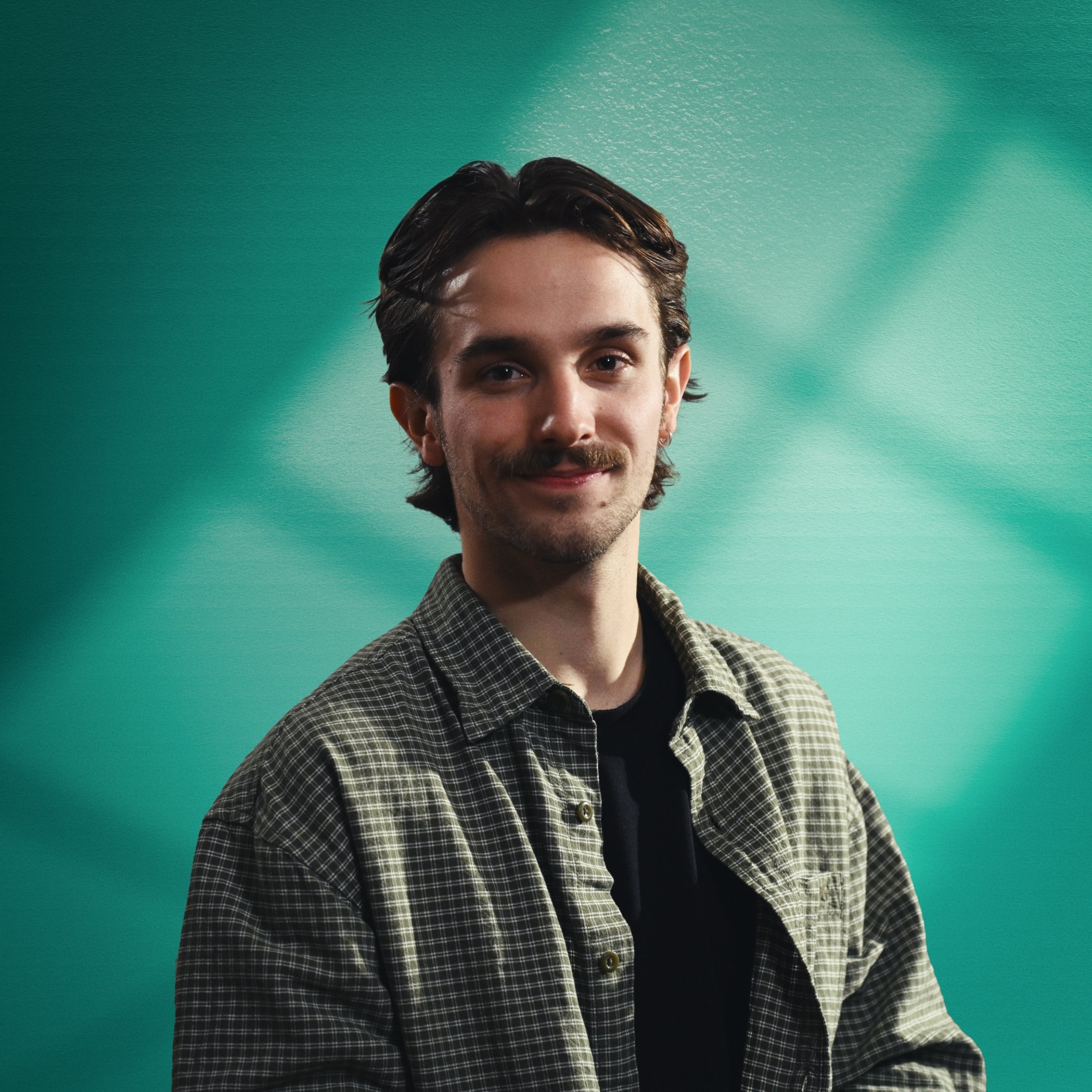 A young man with dark hair and facial hair, wearing a checkered shirt, smiling against a turquoise background. This is Mad Heron's Executive Producer, Lewis Kerr. Lewis is one of Mad Heron's Founding Members.