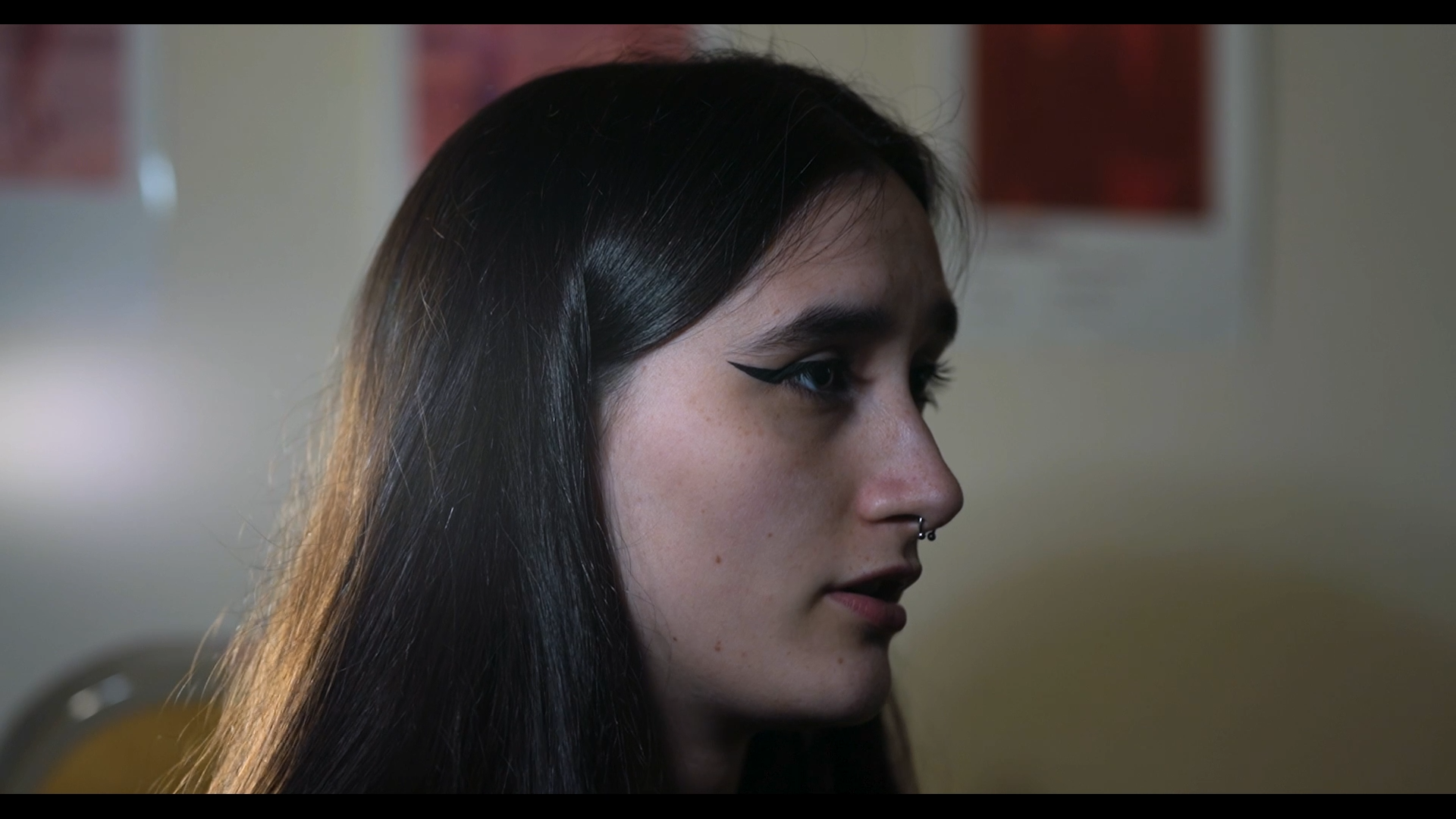Close-up side profile of a young woman with long dark hair, a septum piercing, winged eyeliner makeup, and freckles, in an indoor setting.