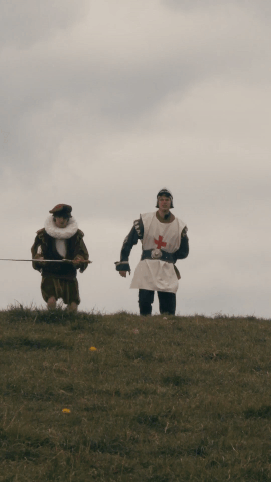 Two people in medieval costumes walking on a grassy hill under a cloudy sky with a dragon flying over head. This is a commercial/advertisement for the mobile phone game, AFK journey.