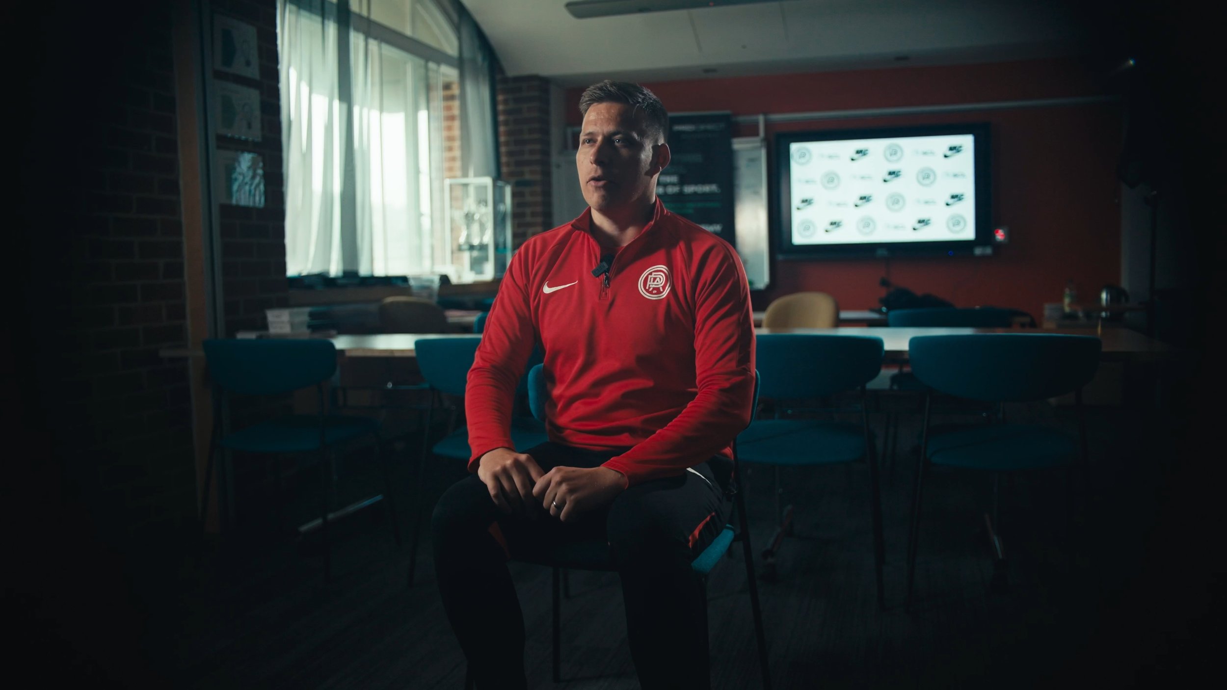 Man wearing a red Nike sports jacket with a logo, sitting in a room with chairs and a large screen in the background.