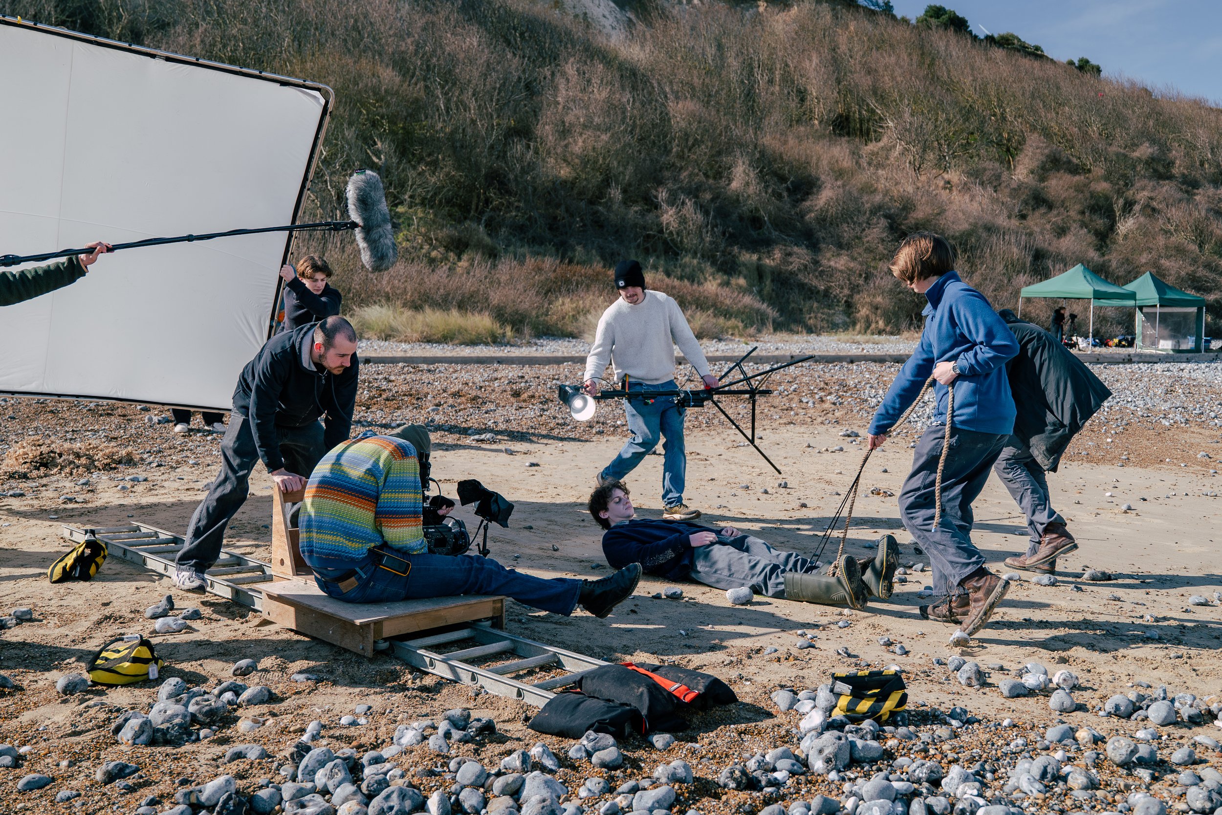 Film crew members filming a scene on a rocky beach, with a person lying on the ground, surrounded by equipment, with sand, rocks, and hillside in the background. This is a behind-the-scenes still from our award-winning short film 'Lure'.