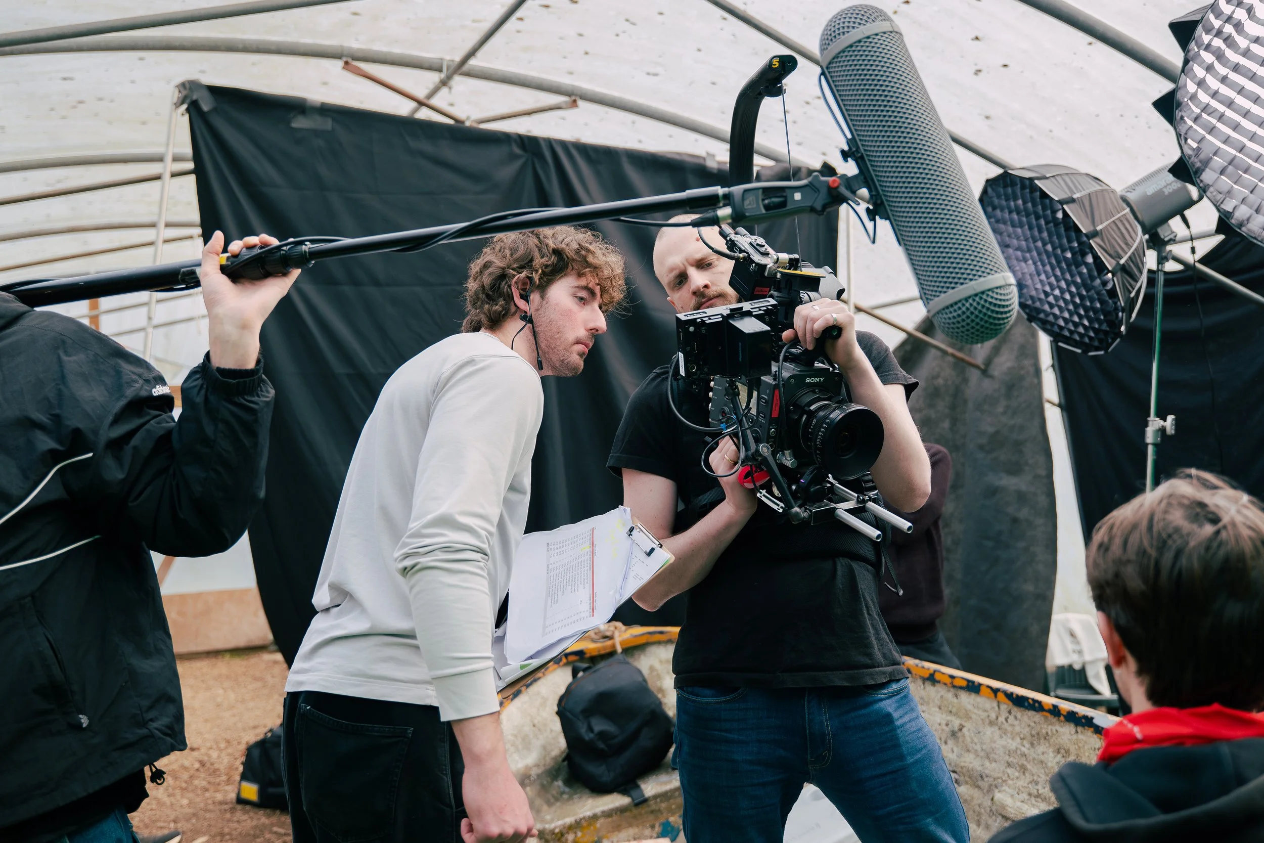 Filmmakers setting up camera and audio equipment inside a polytunnel being used as a green screen studio. Production. Sony Venice Camera. Easy Rig. Boom Microphone. Nanlite. Amaran. Professional studio lighting.