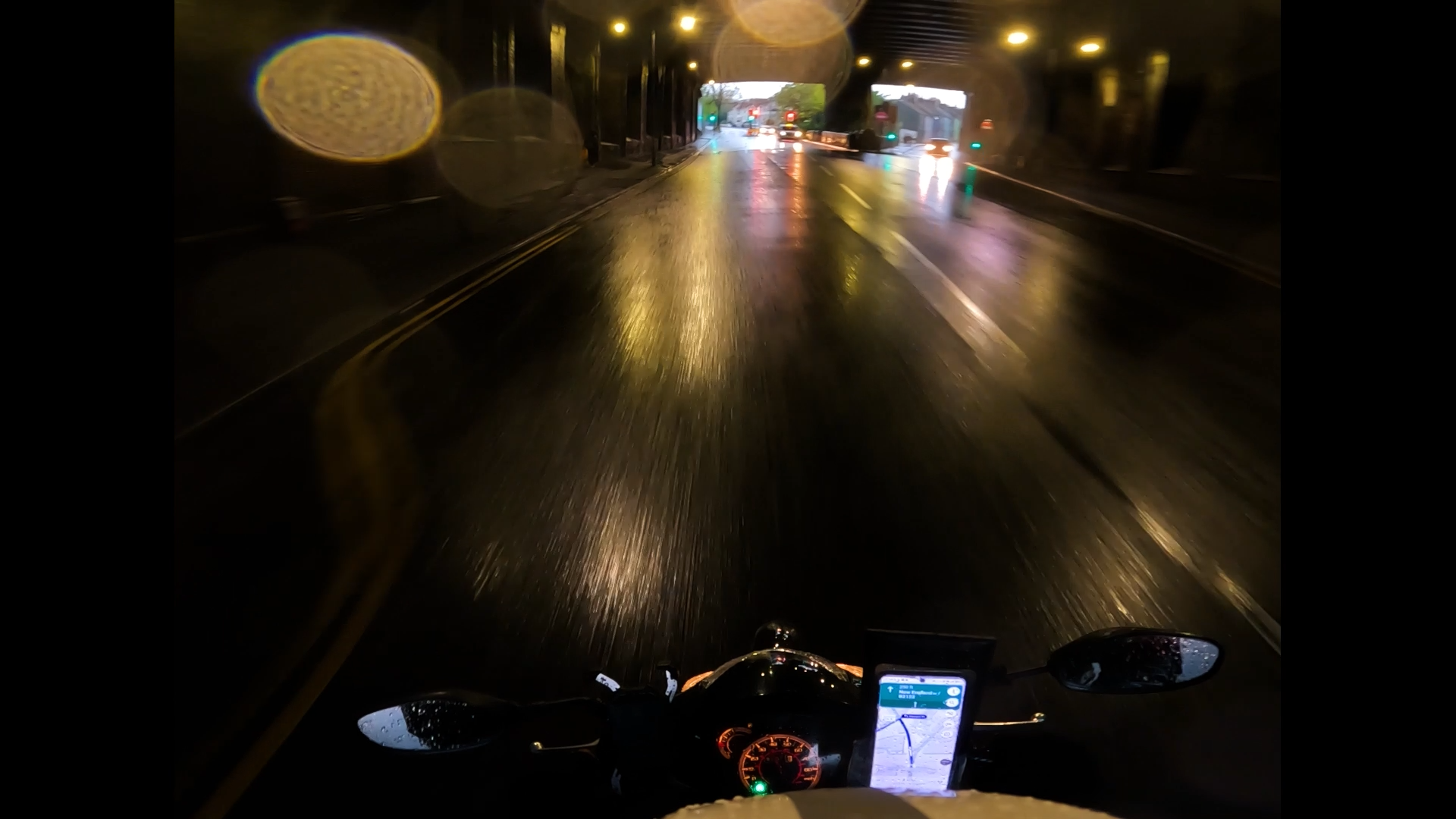 View from a motorcycle rider's perspective on a wet road at night with rain droplets on the camera lens, illuminated by streetlights and vehicle headlights, with a GPS device mounted on the handlebar.
