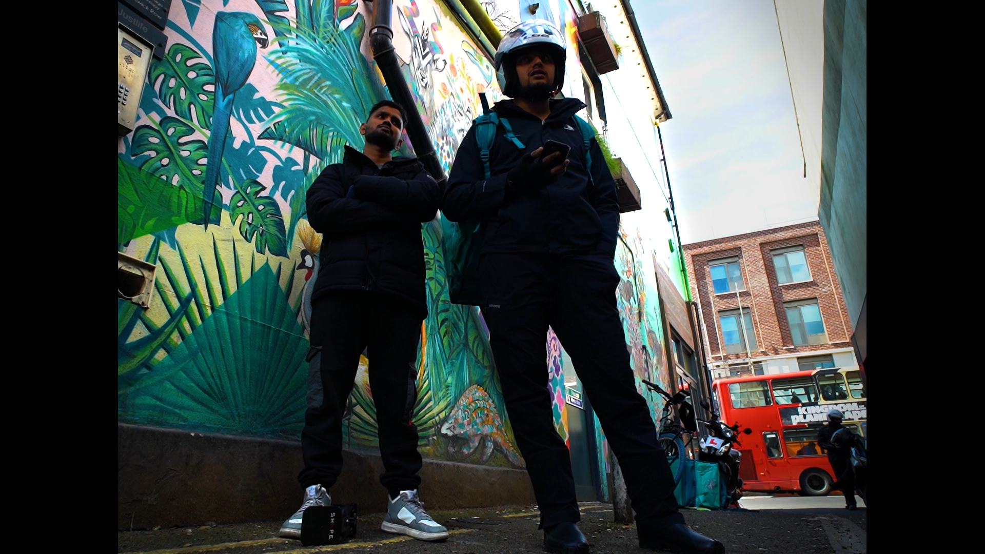Two men standing against a colorful graffiti-covered wall in an urban street scene, with a red double-decker bus in the background.