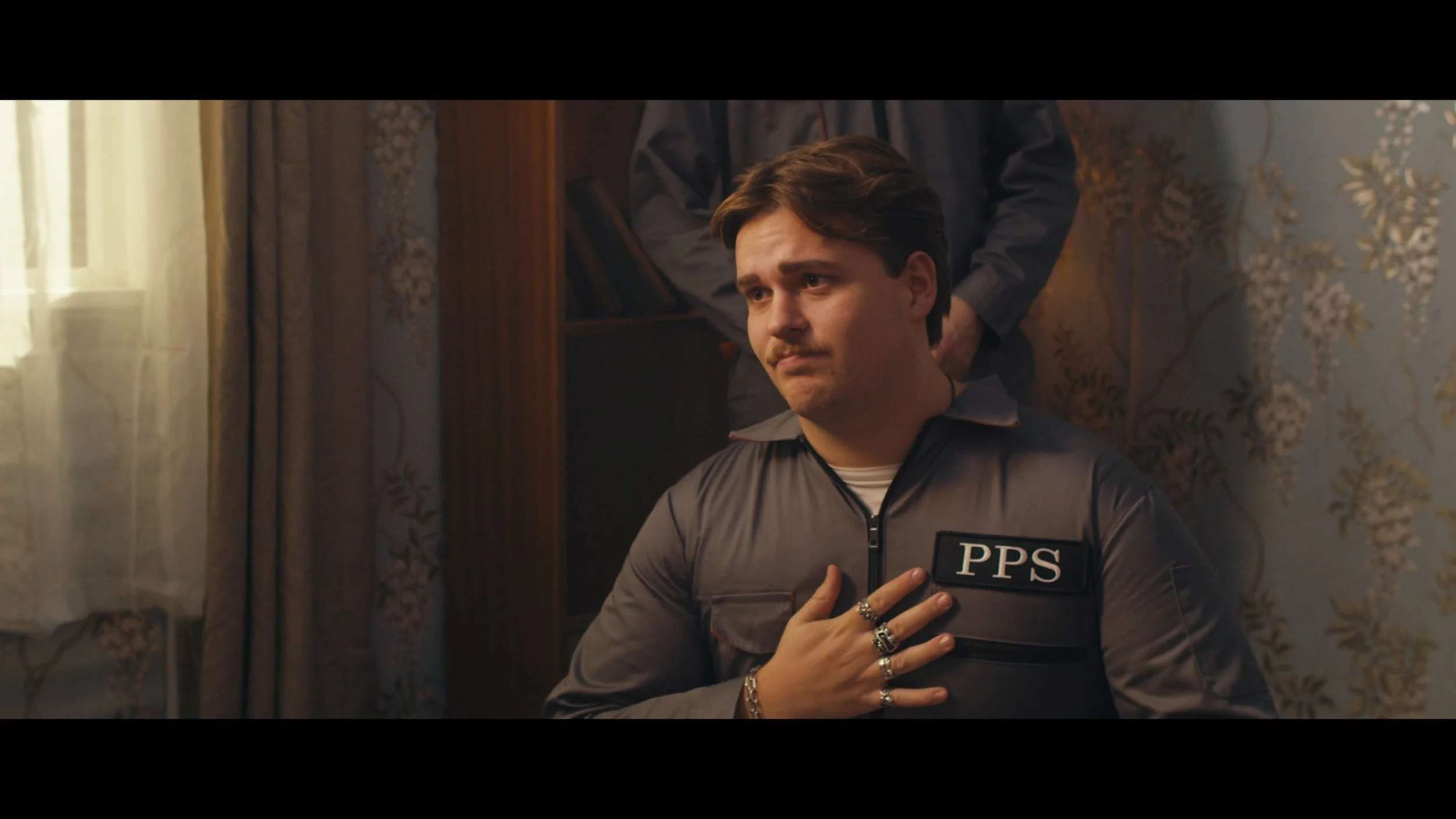 A young man dressed as a police officer with a 'PPS' badge on his uniform, standing in a room with floral wallpaper and curtains, with his right hand over his heart.