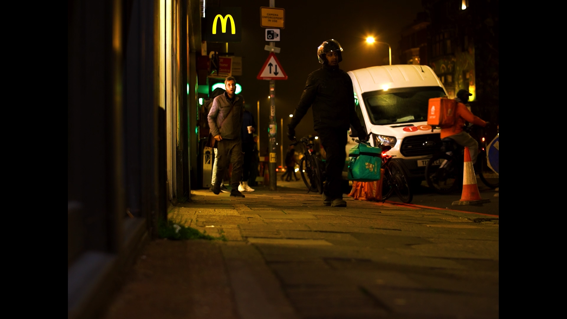 Nighttime urban street scene with people on the sidewalk, a delivery person on a bicycle, a white van, a McDonald's sign illuminated, and streetlights casting warm glow.