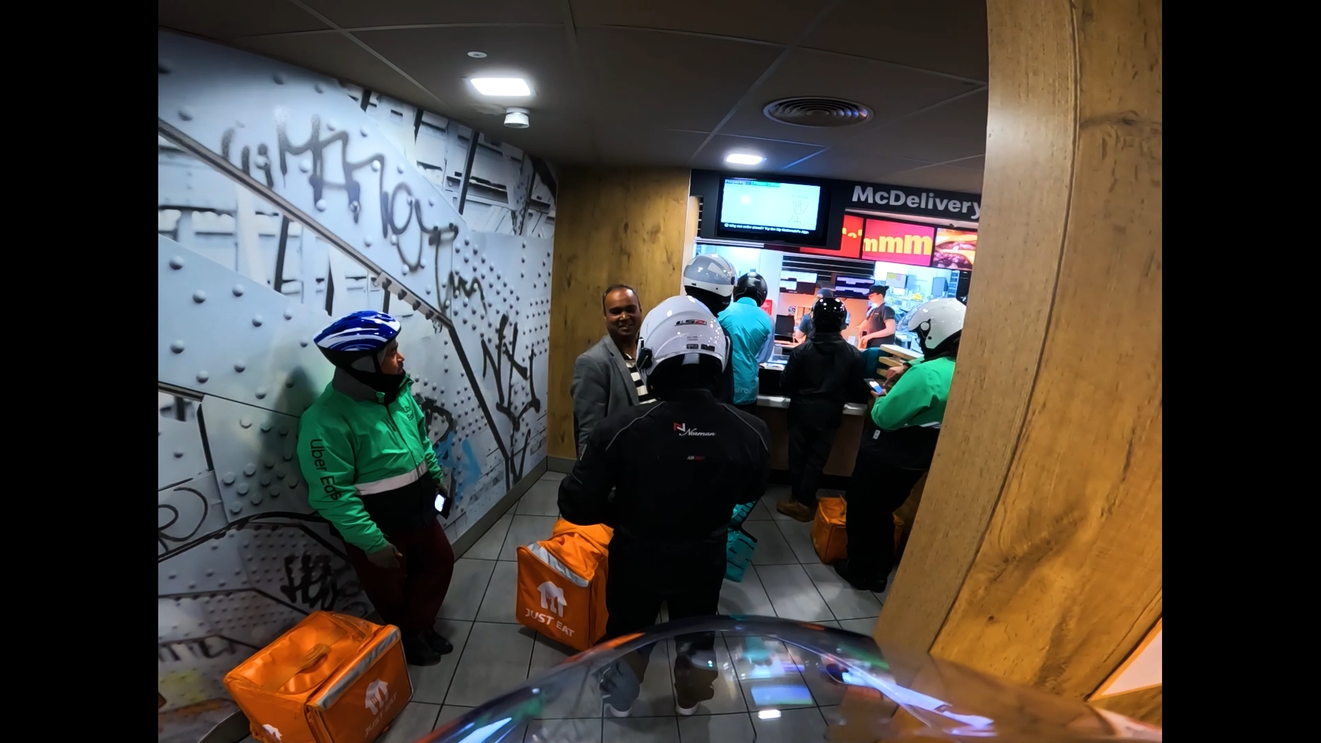 Fast food restaurant interior with people waiting and looking at digital screens, some wearing helmets and holding delivery bags, graffiti wall on left, wooden panel on right.
