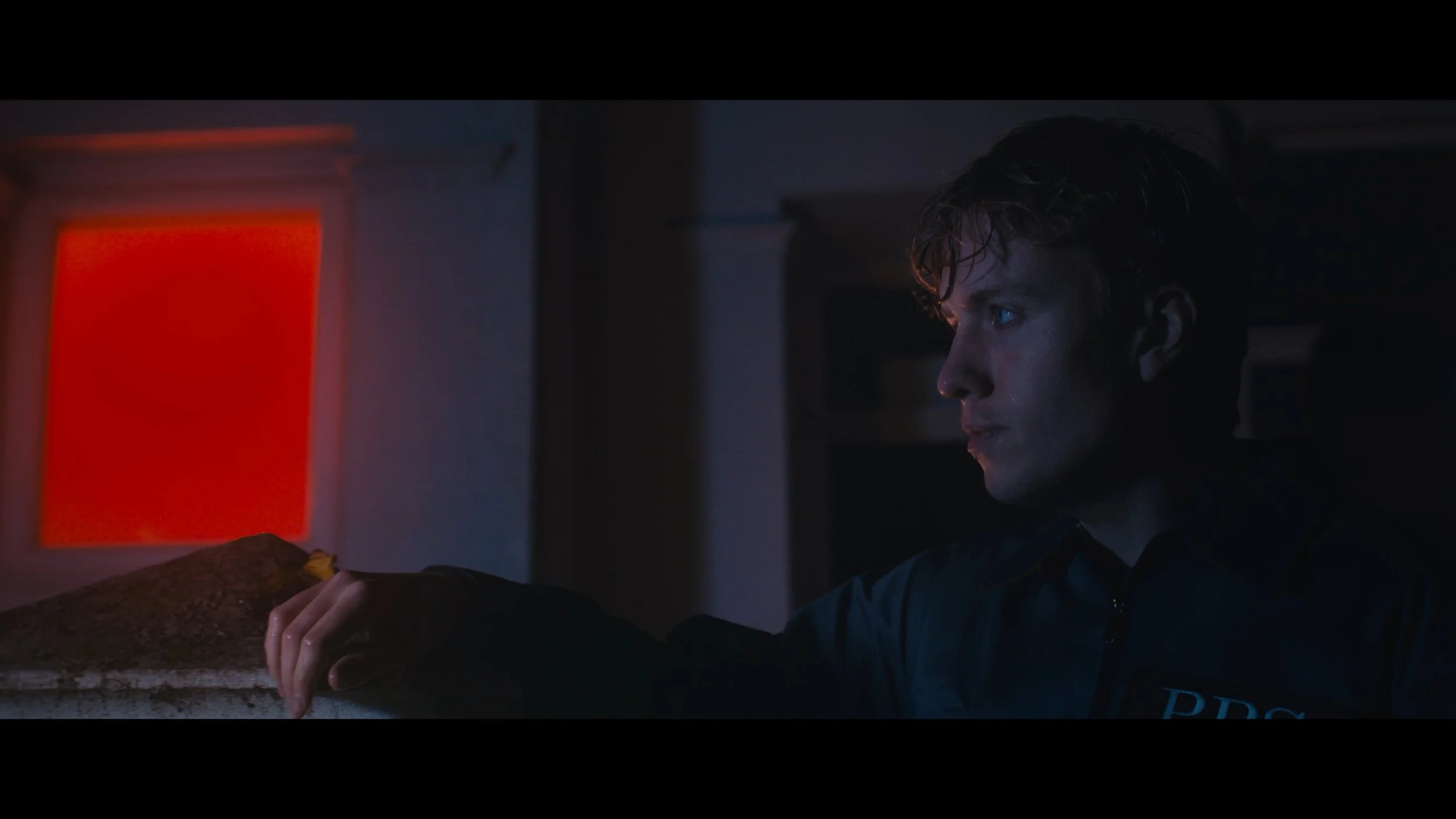 A young man with wet hair and a serious expression extends his arm, with a mound of dirt or soil on his hand, in a dimly lit room with a red illuminated window or panel in the background.