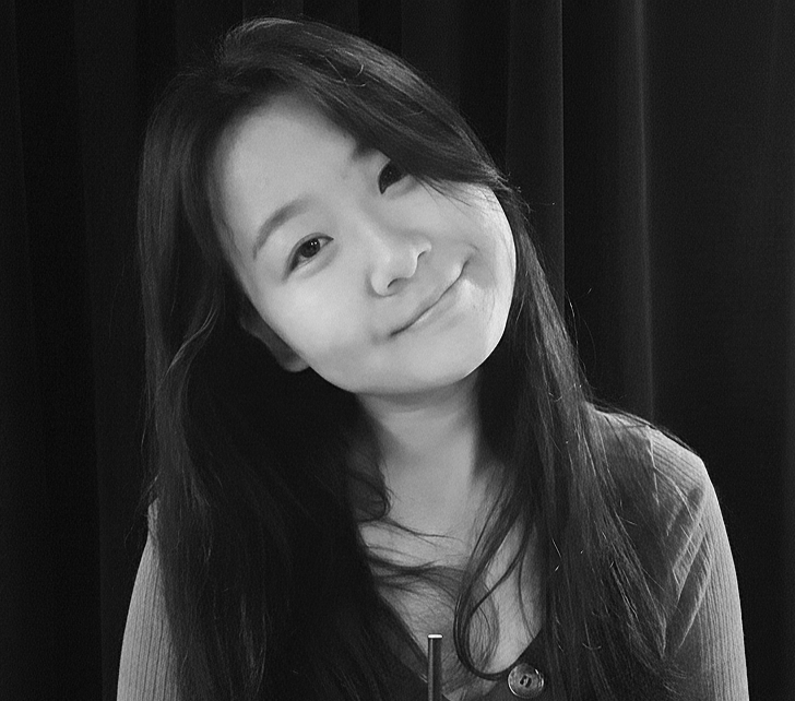 A young woman with long dark hair smiling slightly and tilting her head, in front of a dark curtain.