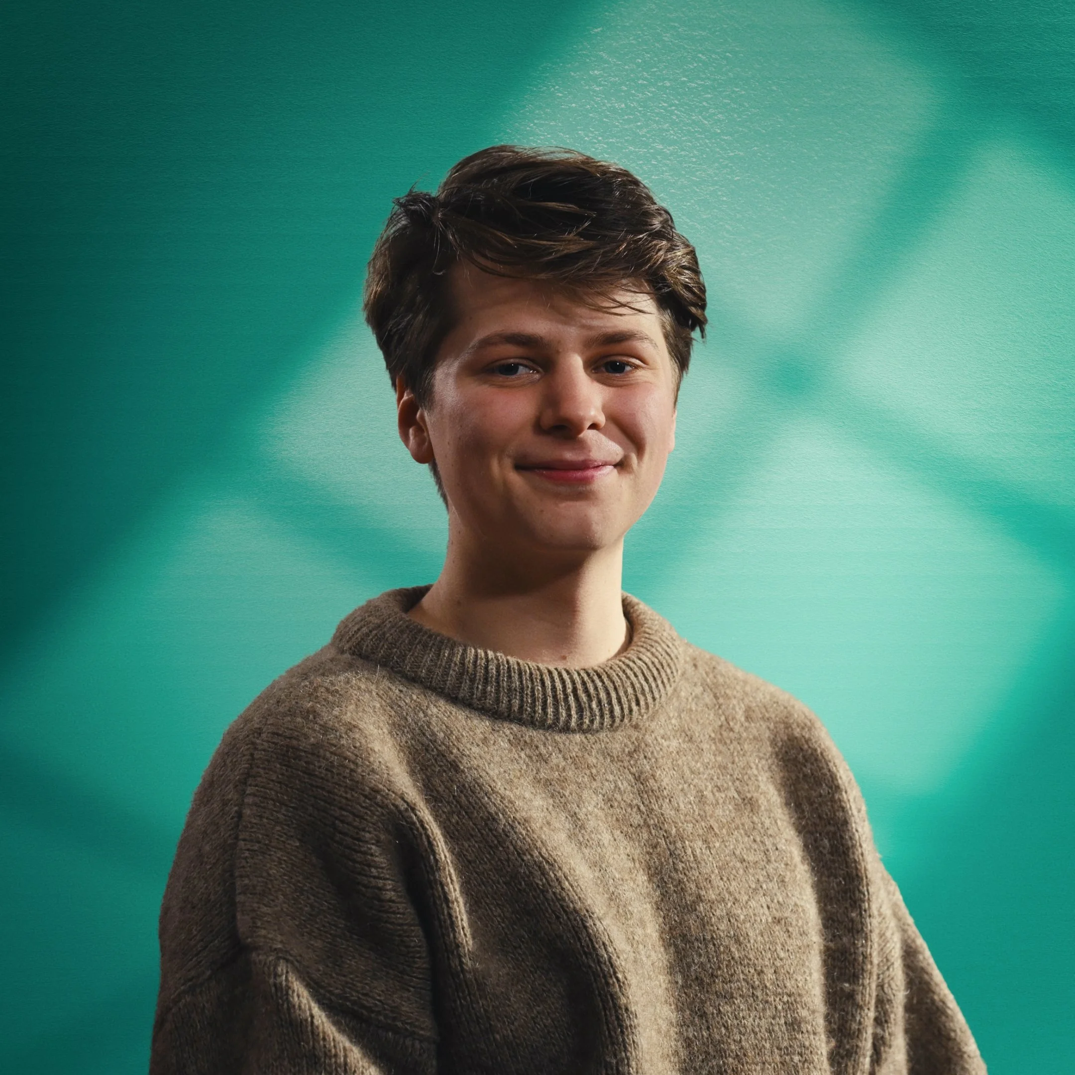 Young person with short brown hair smiling in front of a teal gradient background, wearing a beige knitted sweater. This is Mad Heron's Creative Director Jasper Pagan. Jasper is one of Mad Heron's Founding Members.