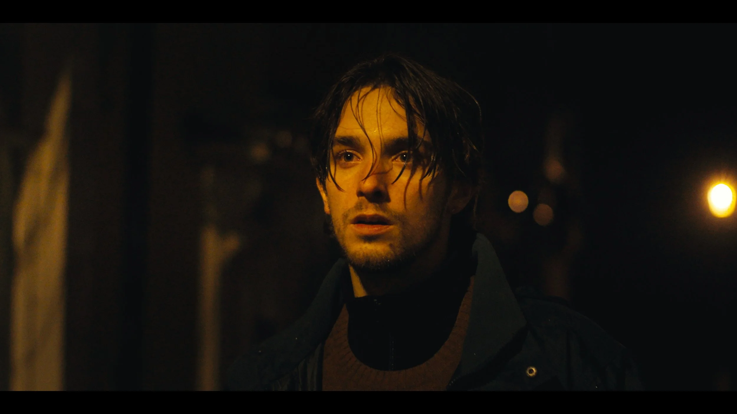 A young man with wet, tousled dark hair and a serious expression, standing in a dimly lit outdoor setting at night.