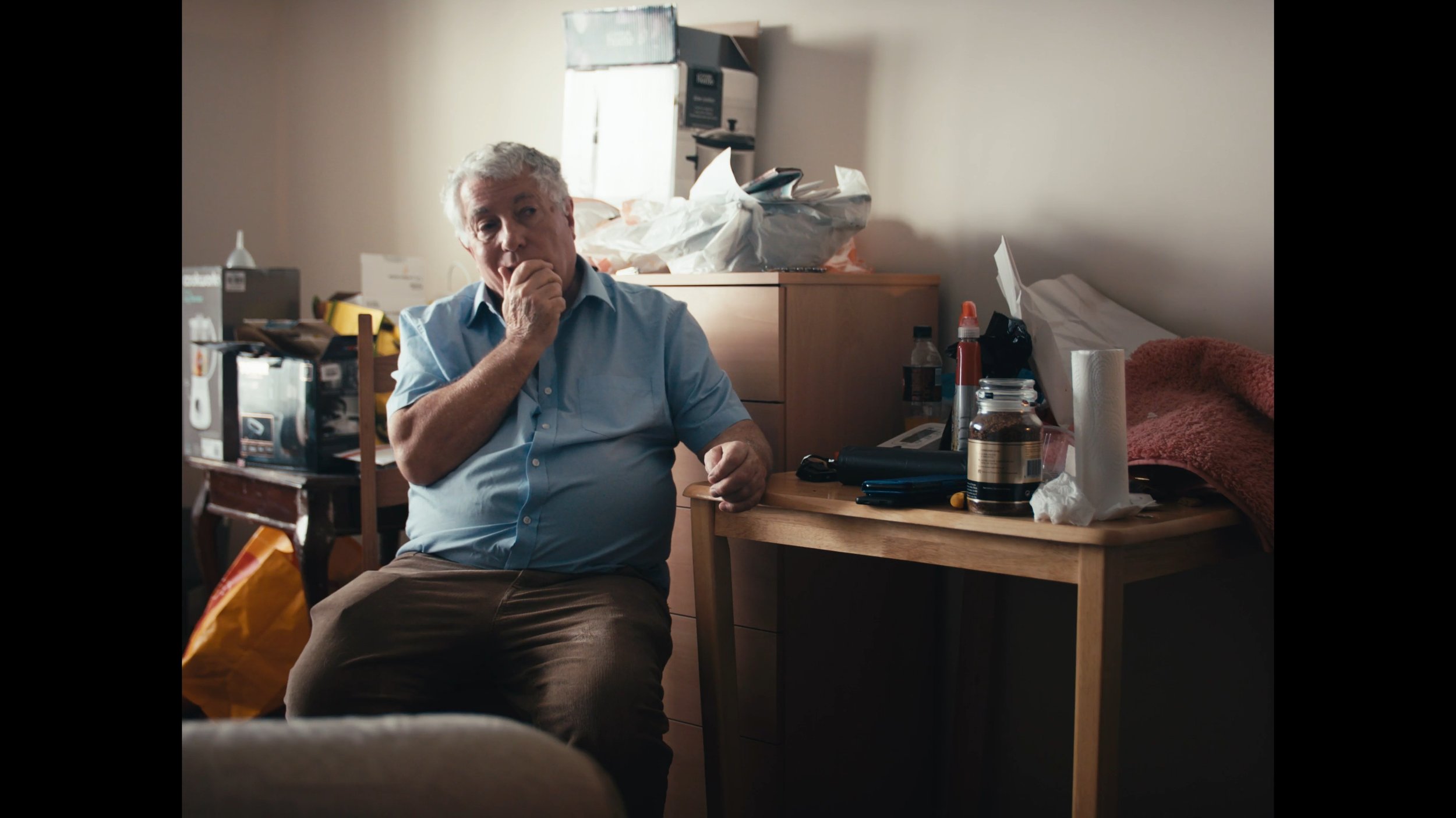 An elderly man with gray hair wearing a light blue shirt, sitting in a cluttered room with various objects such as bottles, paper towels, and bags around him, looking contemplative.