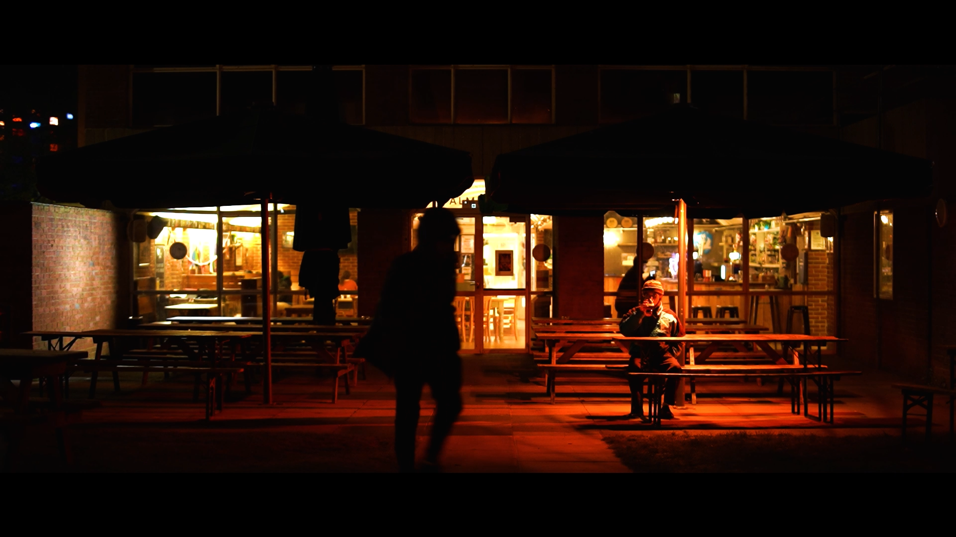 Nighttime scene outside a brick restaurant with outdoor seating under large umbrellas, illuminated from inside. One person is sitting alone at a table, smoking. A person is walking past the restaurant, and other people are visible inside through the windows.