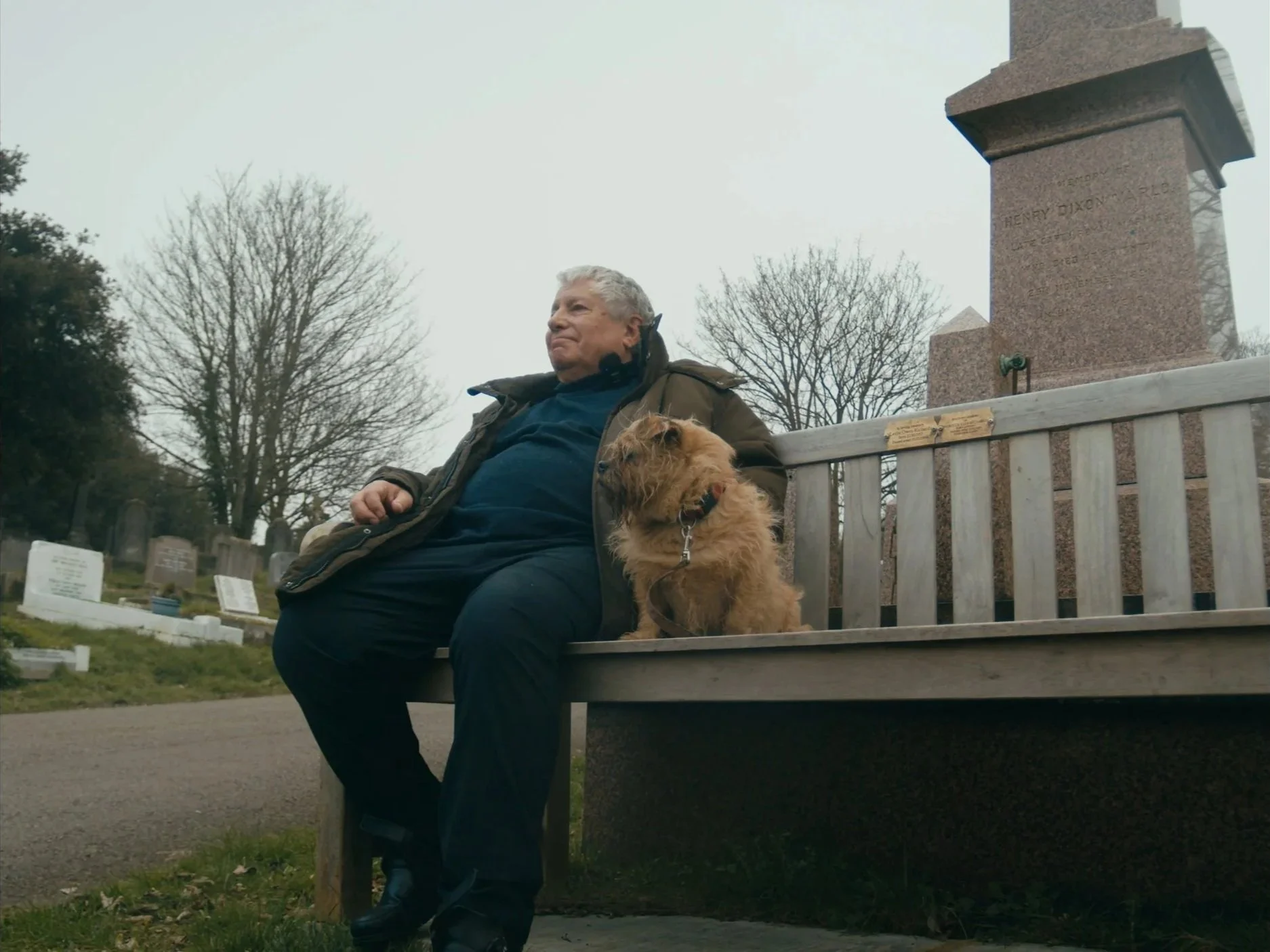 An elderly man sitting on a cemetery bench with a scruffy brown dog, surrounded by tombstones and leafless trees on a cloudy day. Documentary.