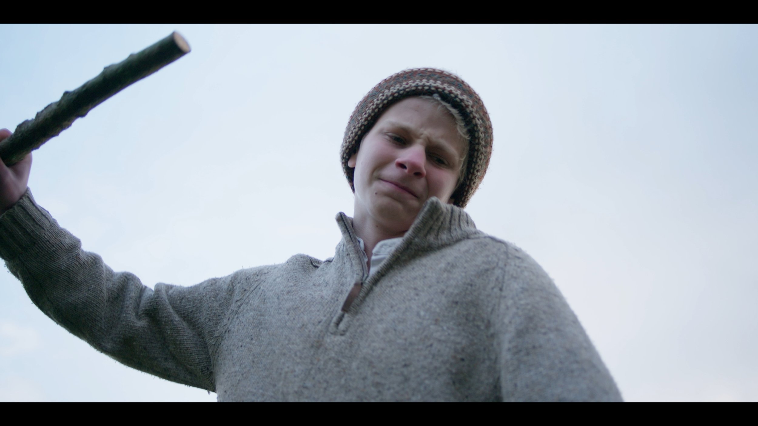 A young boy in a knit cap and gray sweater holding a stick, looking down at the camera with a serious expression against a cloudy sky.
