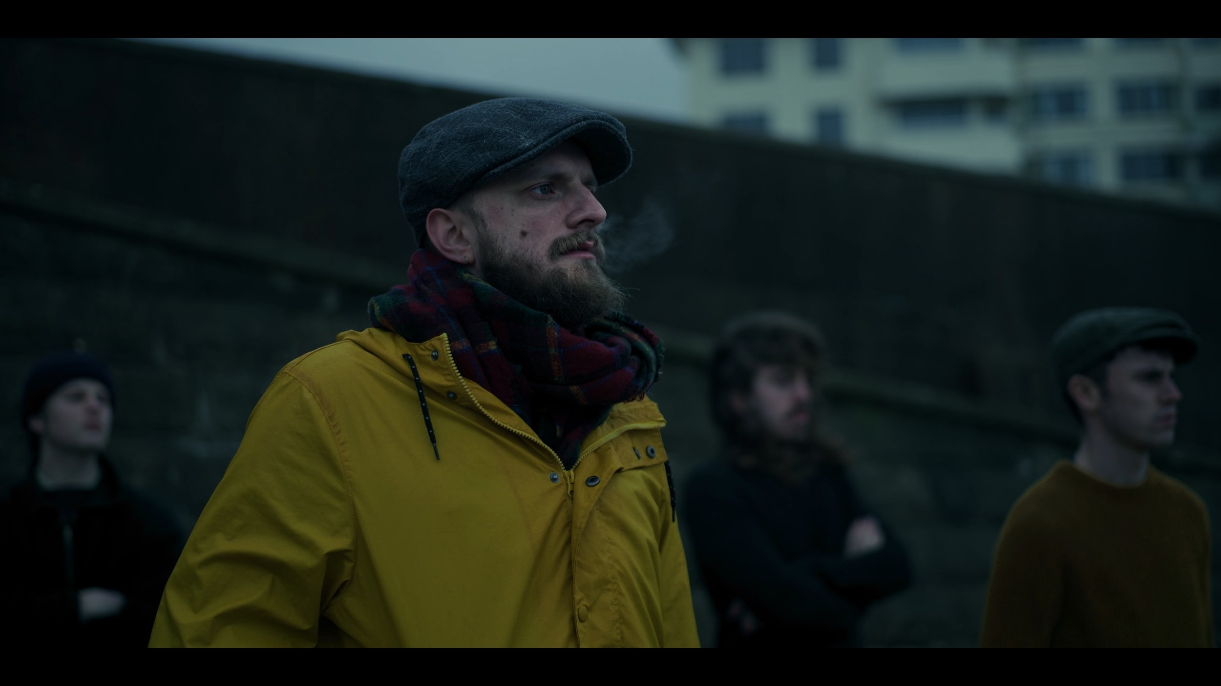 Four people standing outdoors near a wall, with a city building in the background. The man in the foreground wears a yellow jacket, a gray flat cap, and a plaid scarf, looking serious. The other three people are blurred, dressed in dark clothing.