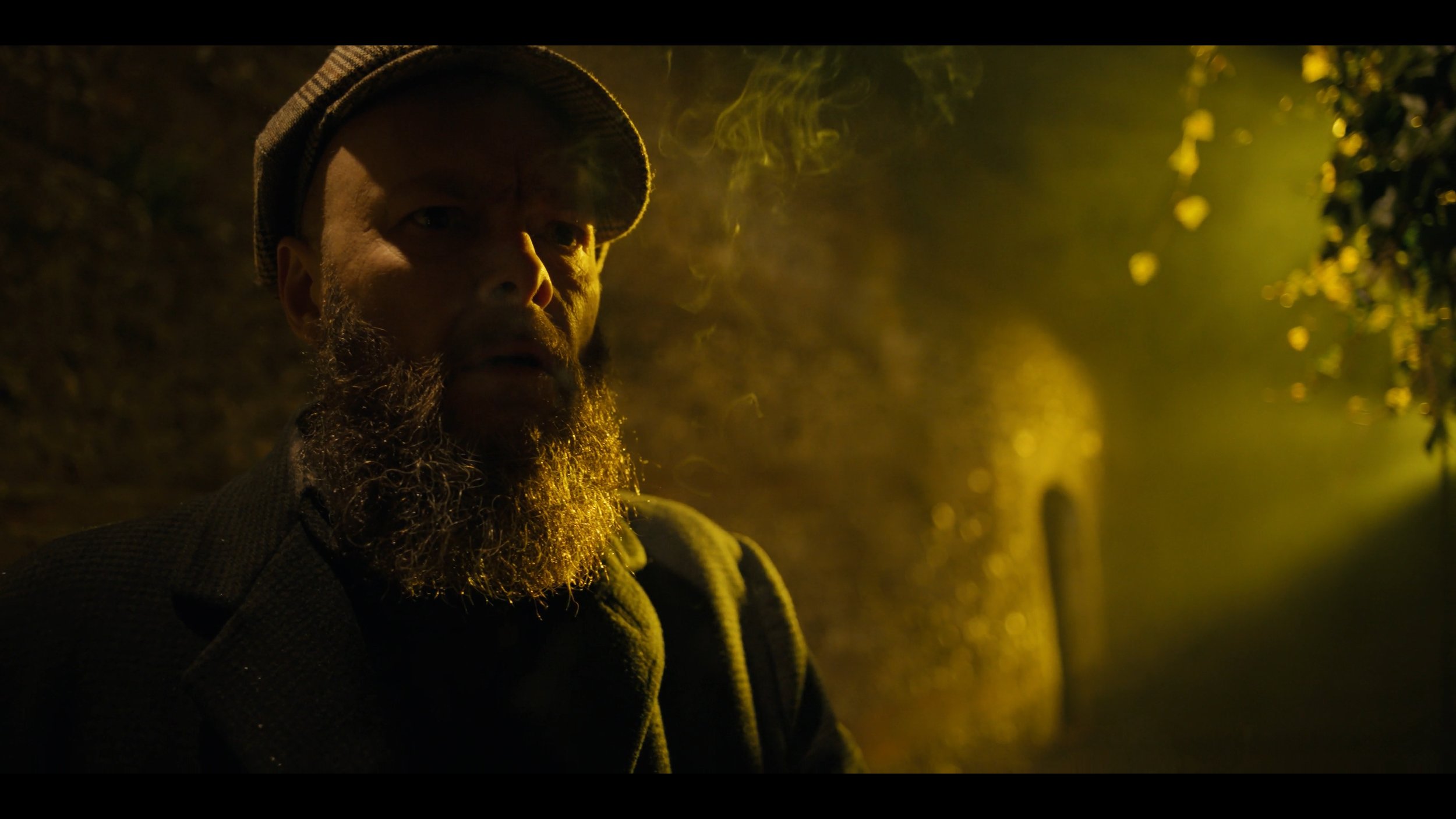 A man with a beard and a cap sitting outdoors at night, illuminated by a warm yellow light, with a blurred background.