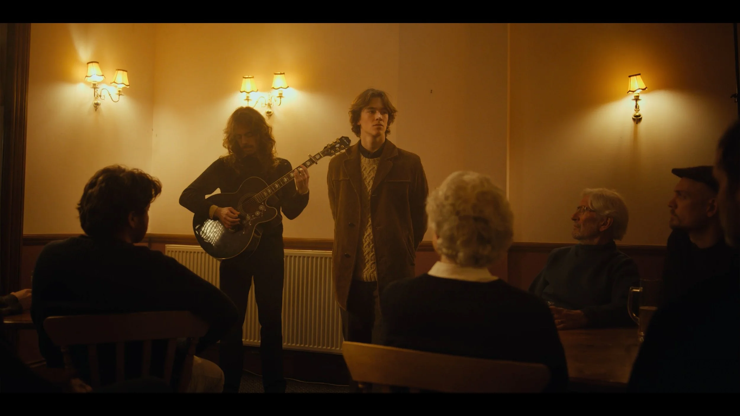 A young man stands in a dimly lit room with warm lighting, watching a musician play guitar. The scene features five people, including the young man, seated and facing the musician, who is performing in front of a wall with two wall-mounted lamps, creating an intimate setting.