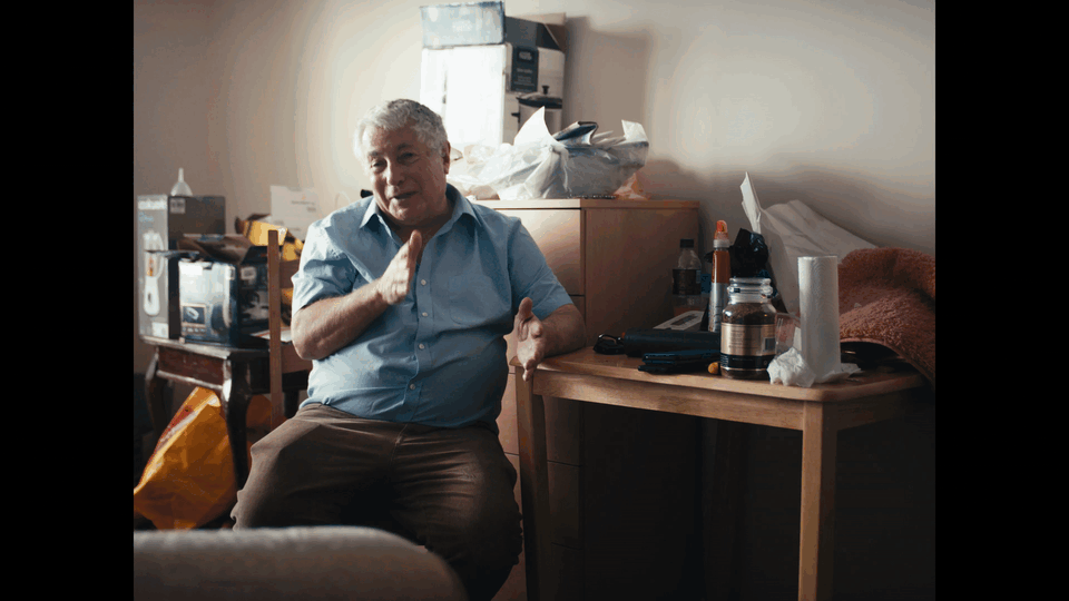 An elderly man sitting on a bed in a cluttered room, gesturing with one hand, with various objects like bottles, tissues, and boxes on a small wooden table next to him.