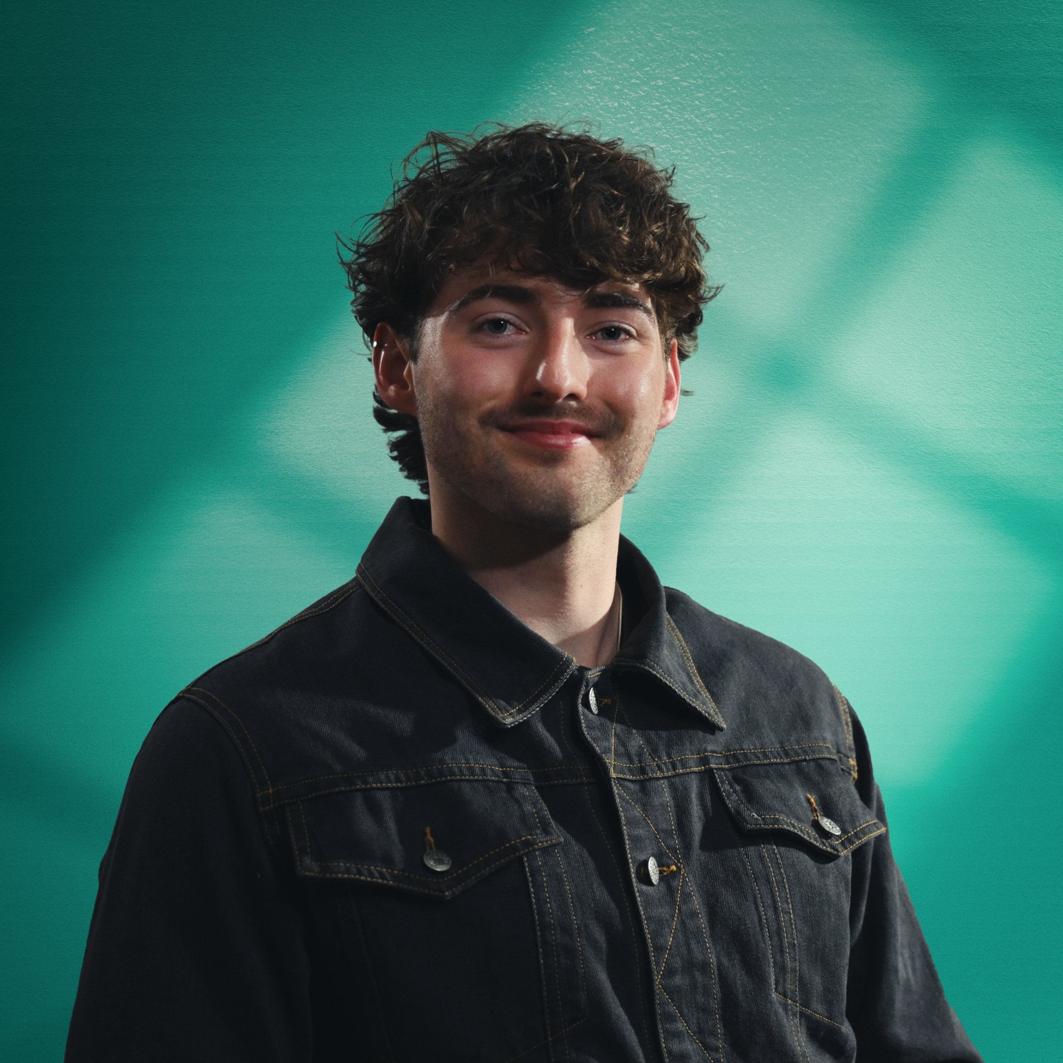 Young man with curly brown hair wearing a black denim jacket, smiling against a teal background. This is Mad Heron's Head of Production, Joe Shale. Joe is one of Mad Heron's Founding Members.