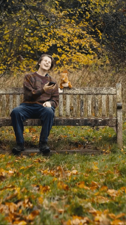 A person sitting on a park bench with autumn leaves on the ground, laughing and holding a phone, with a stuffed animal on the bench, and colorful fall trees in the background.