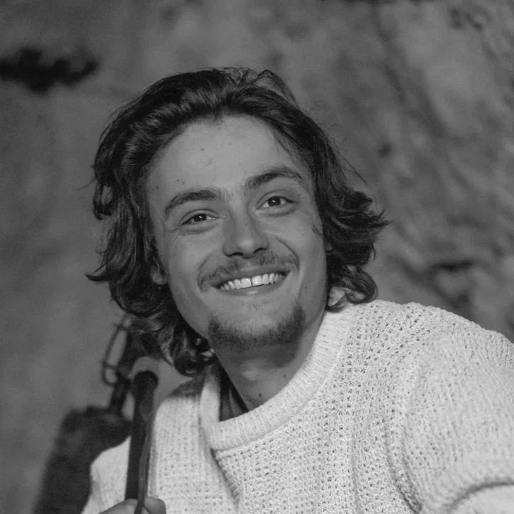 A young man with wavy hair and a beard smiling, wearing a knit sweater, in front of a rocky background.