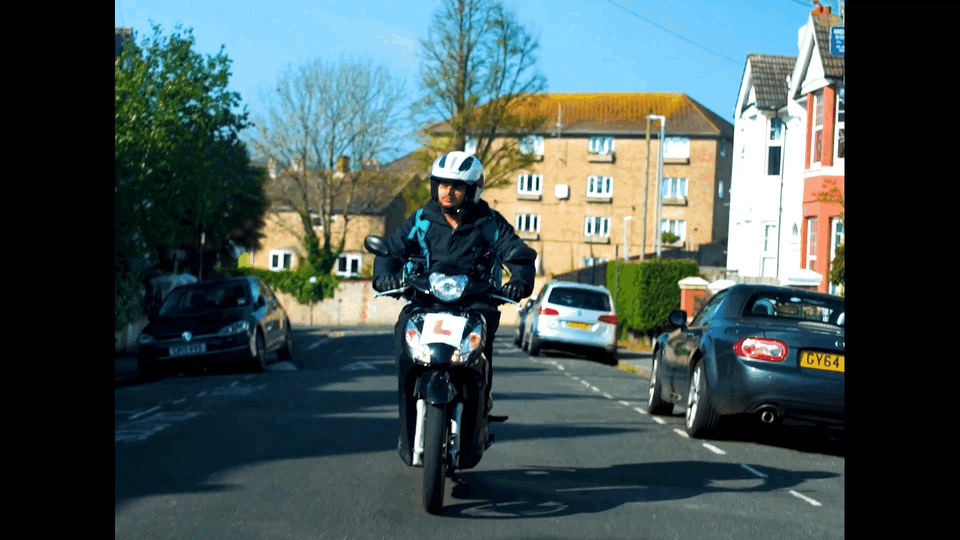 A man riding a motorcycle on a residential street with parked cars and houses in the background.