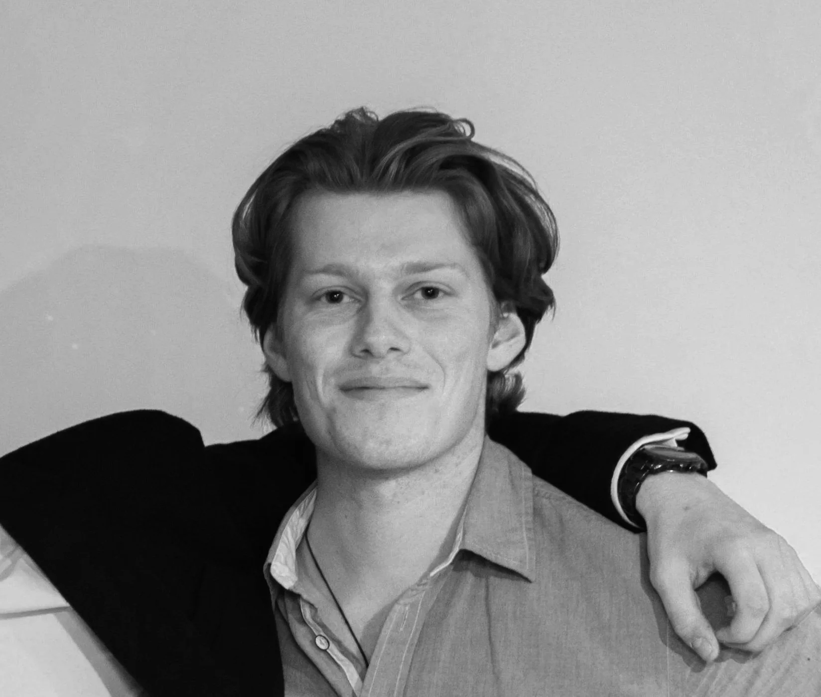 Two young men standing outdoors, one with an arm around the other's shoulder, smiling at the camera. The background shows trees and parts of a house or building.
