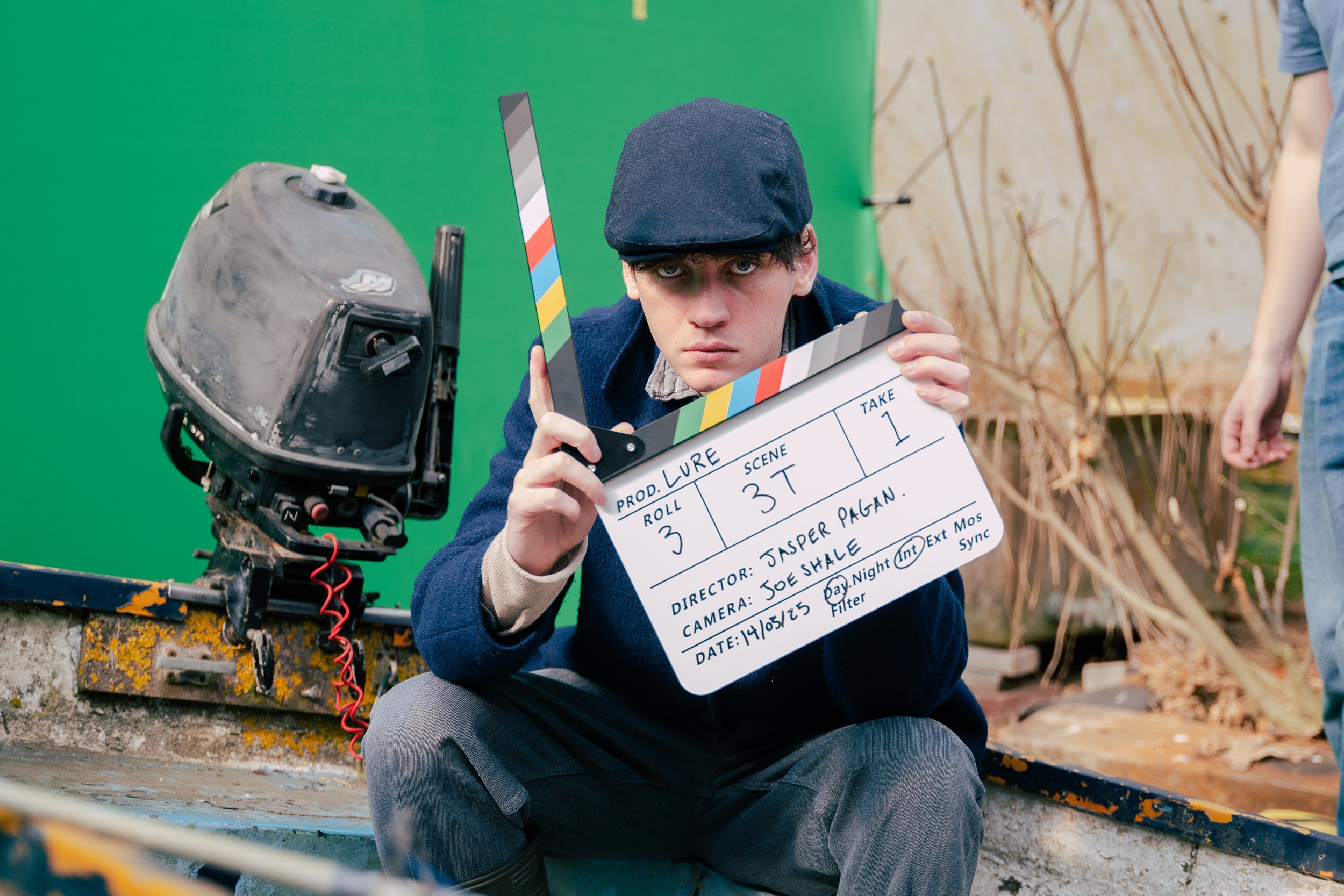 Actor and Model, Reuben Larkin, dressed in vintage clothing holding a film slate on a film set, with a green screen and brown foliage in the background. Narrative Film.