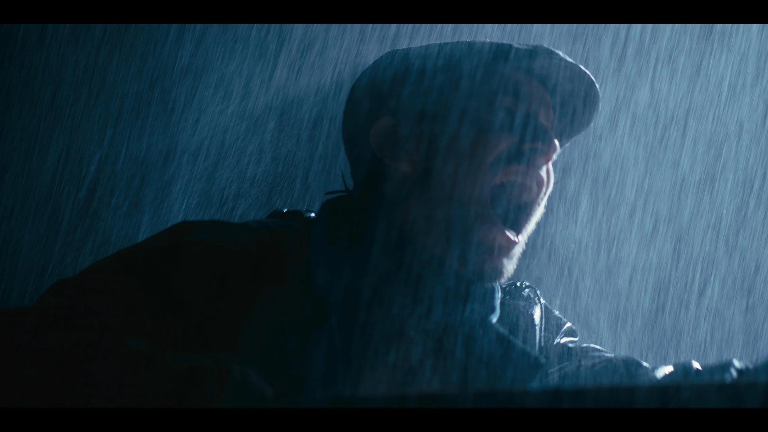 A person wearing a hat and raincoat in the rain, with their mouth open and a joyful expression, illuminated by a blue light.
