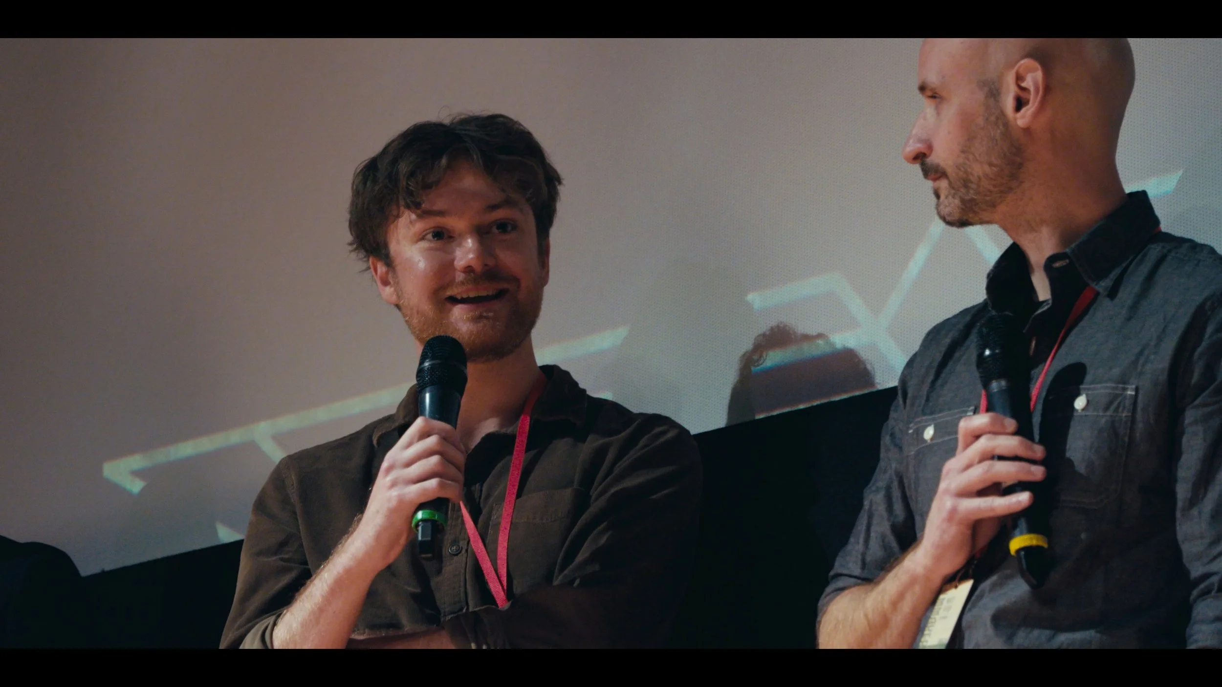 Two men speaking into microphones on a stage with a projected financial chart in the background.