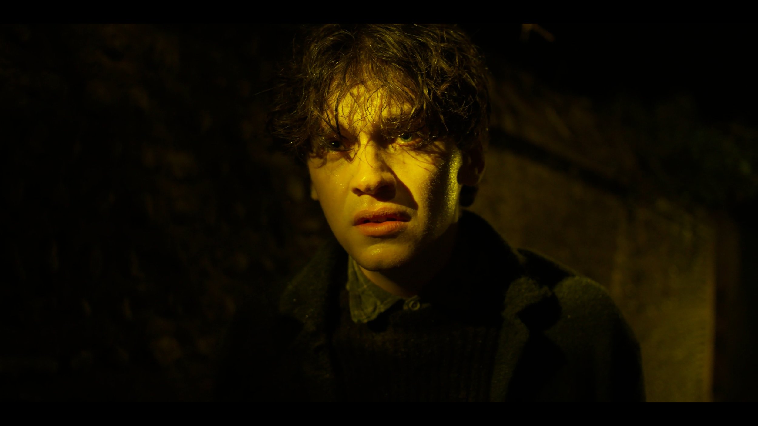 A young man with wet, curly hair stares with a distressed expression in a dimly lit setting.