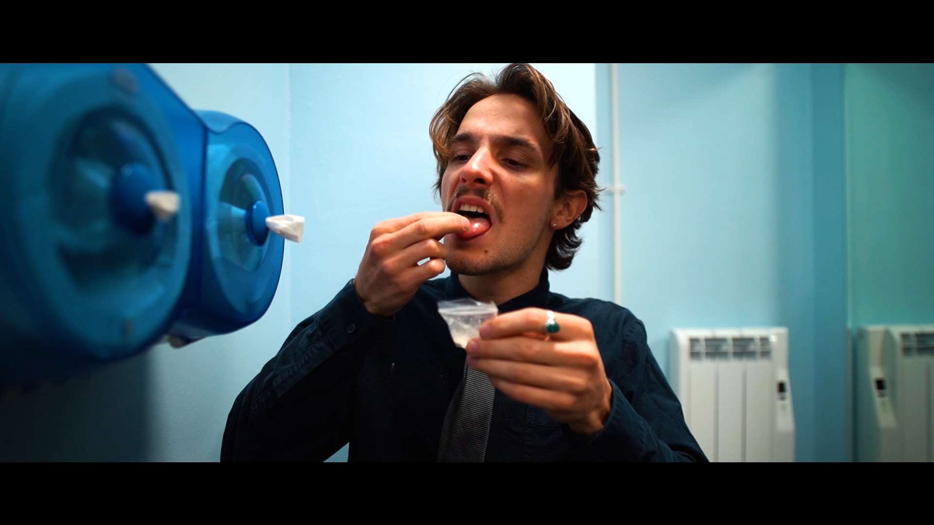 A man with brown hair and earrings is in a bathroom, holding a small plastic bag and taking a pill, with a concerned expression.