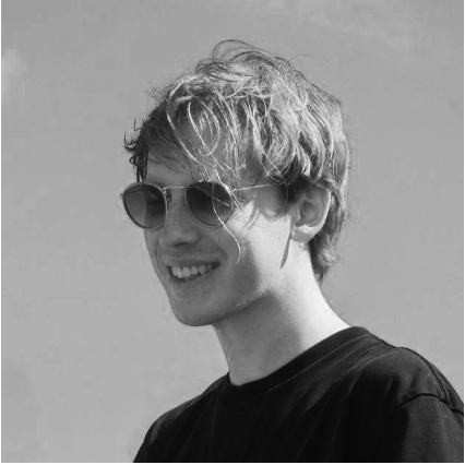 A young man with curly hair wearing sunglasses and a black shirt, smiling outdoors in black and white.