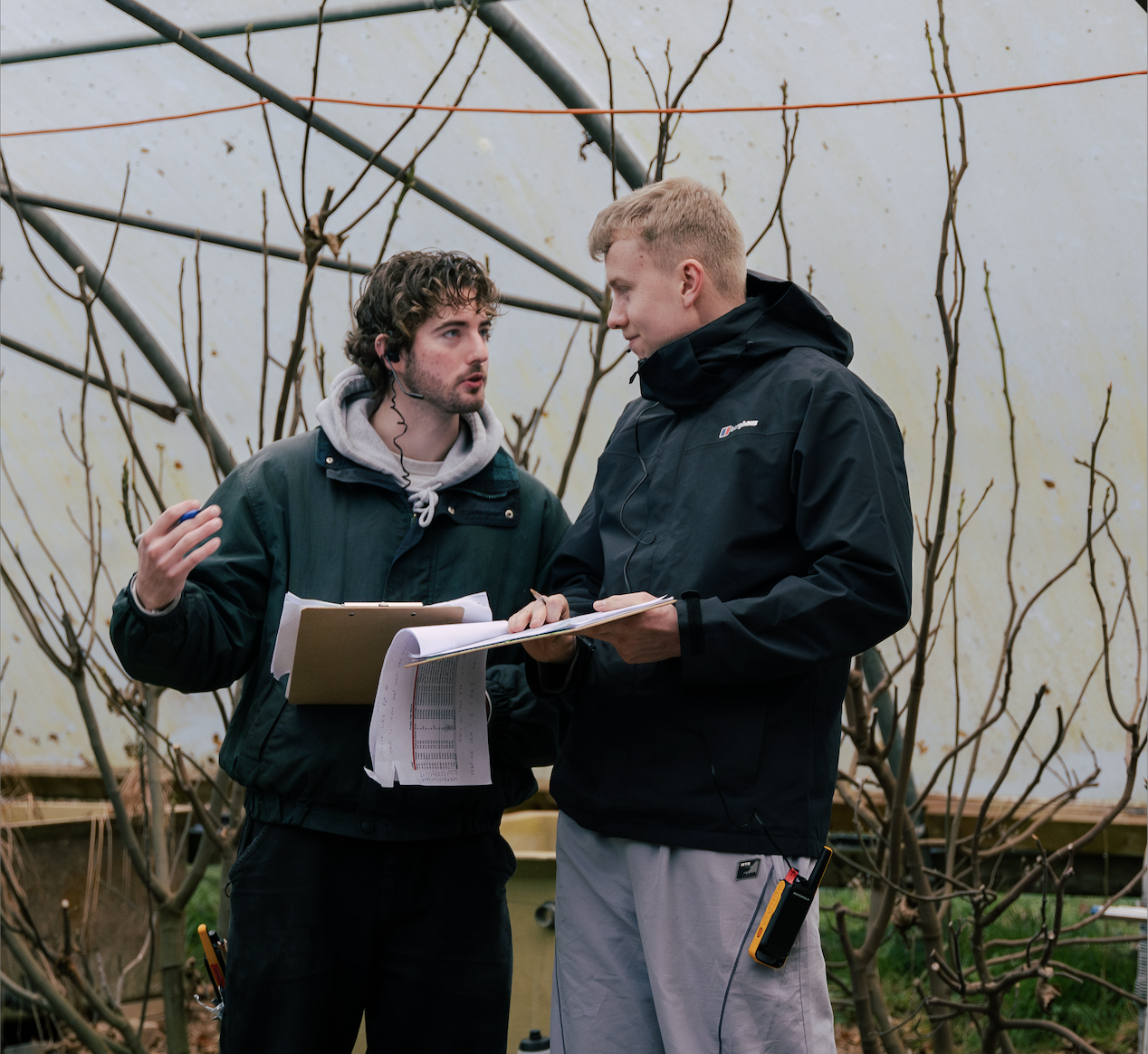 Two men are holding production papers and clipboard on set. One man has curly hair, a headset, and wears a green jacket with a gray hoodie. The other has short blond hair, a black jacket, and a radio clipped to his pocket. Pre-Production.