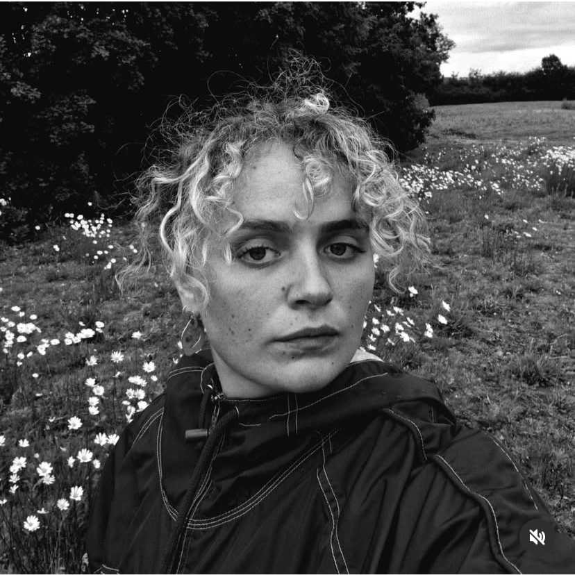 A black-and-white photo of a woman with curly hair, standing outdoors in a field with daisies and trees in the background.